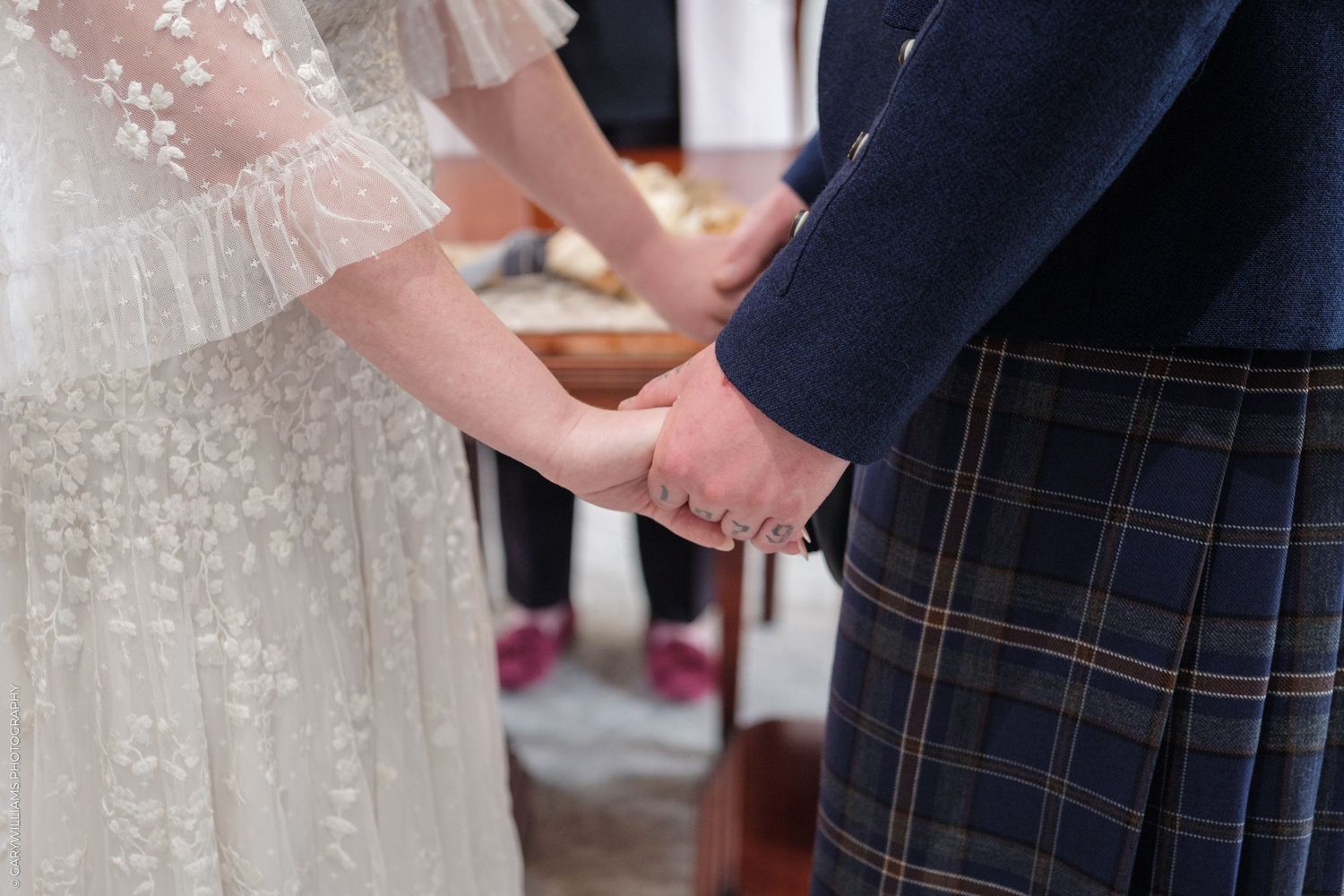 Bride and groom holding hands during wedding ceremony, bride in lace dress, groom in blue tartan kilt.
