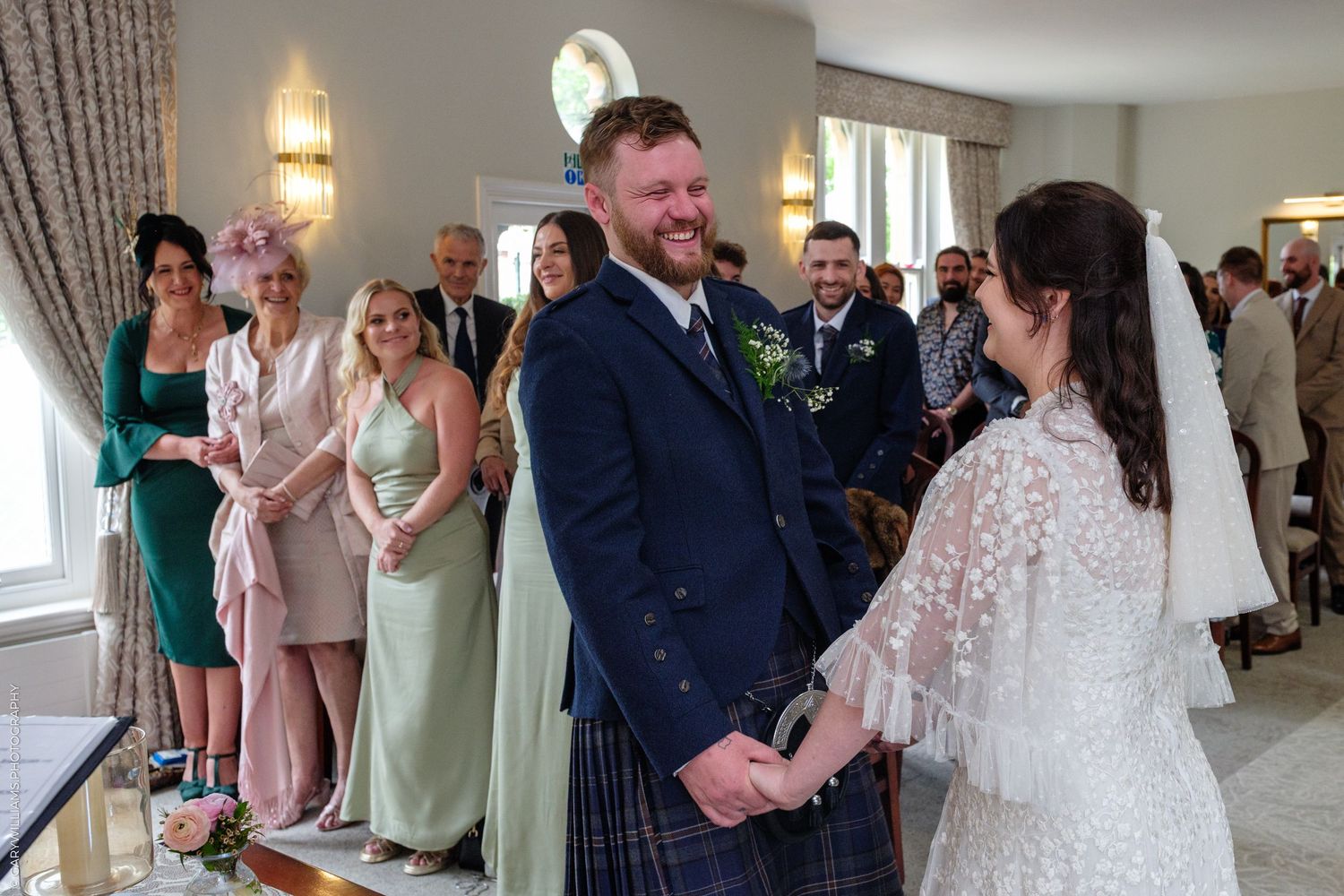 Groom in tartan kilt smiling at bride during indoor wedding ceremony with guests in elegant venue.