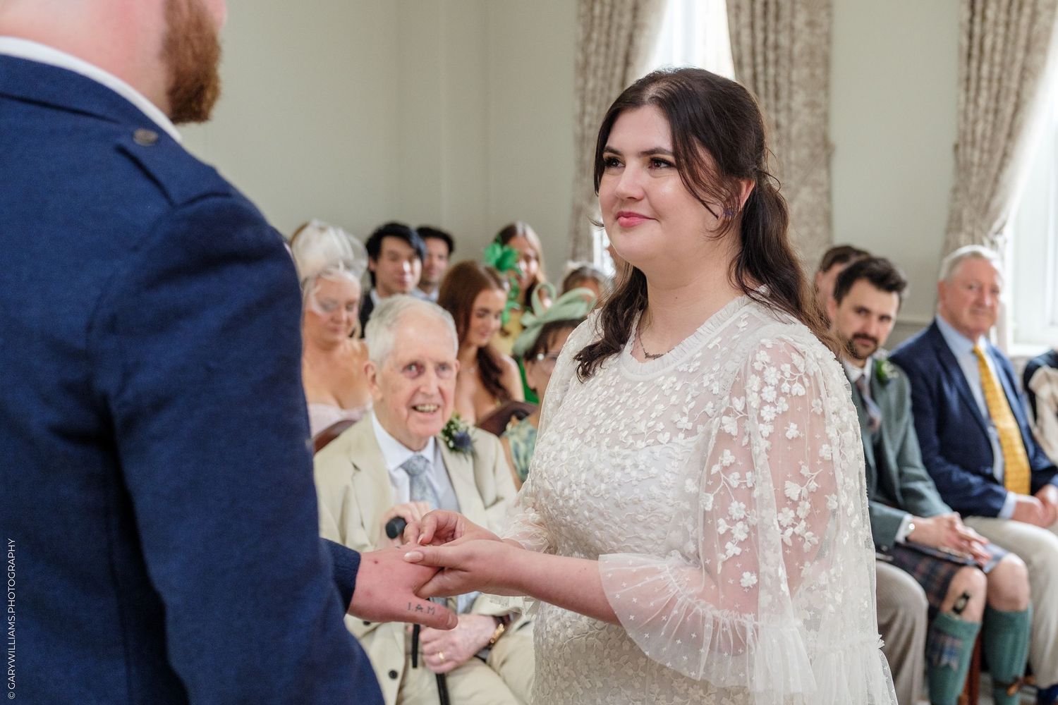 Bride smiling while exchanging rings with groom during an elegant indoor wedding ceremony with seated guests.