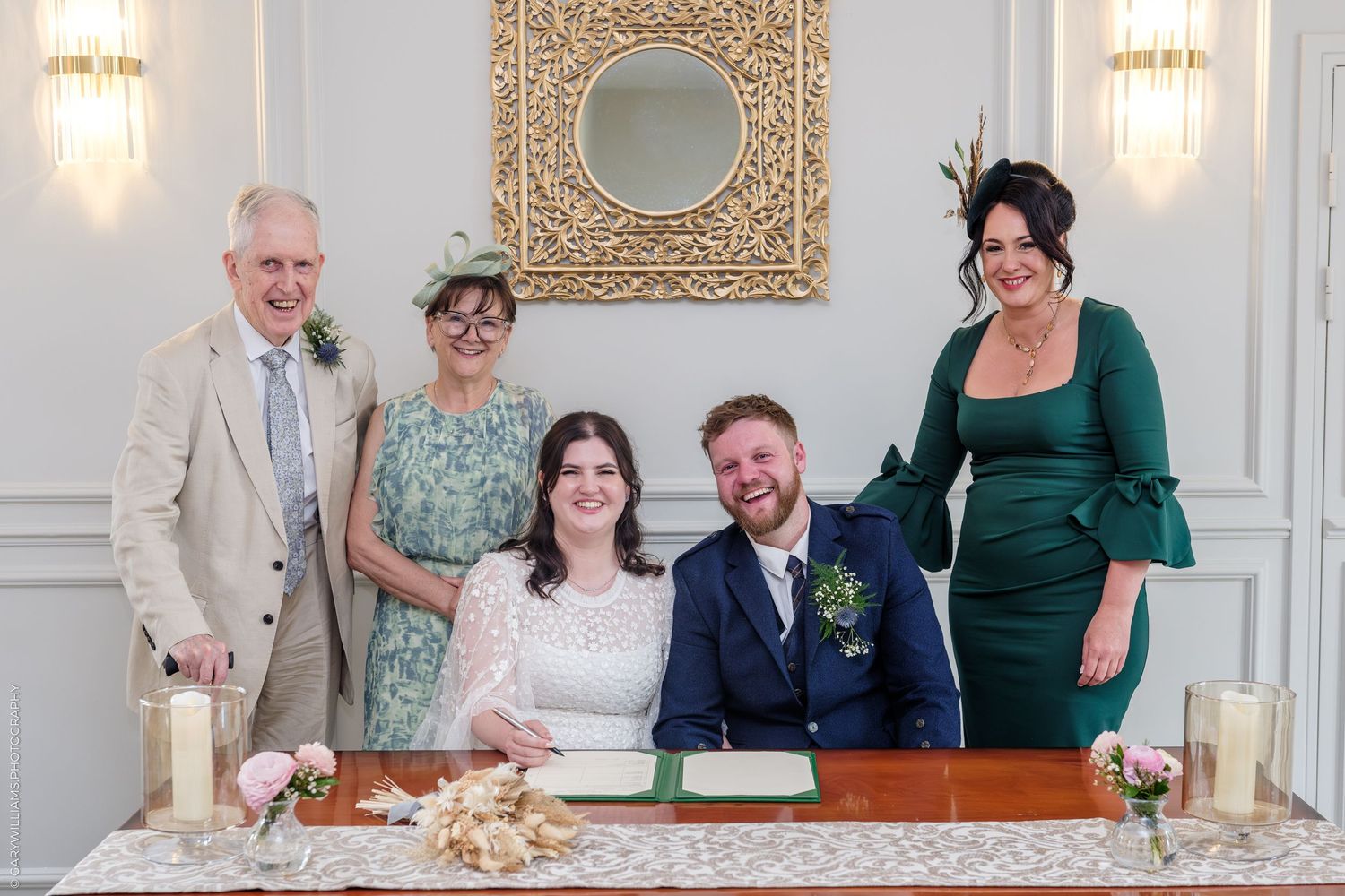 Bride and groom seated at signing table with two female and one male guest at elegant wedding ceremony.