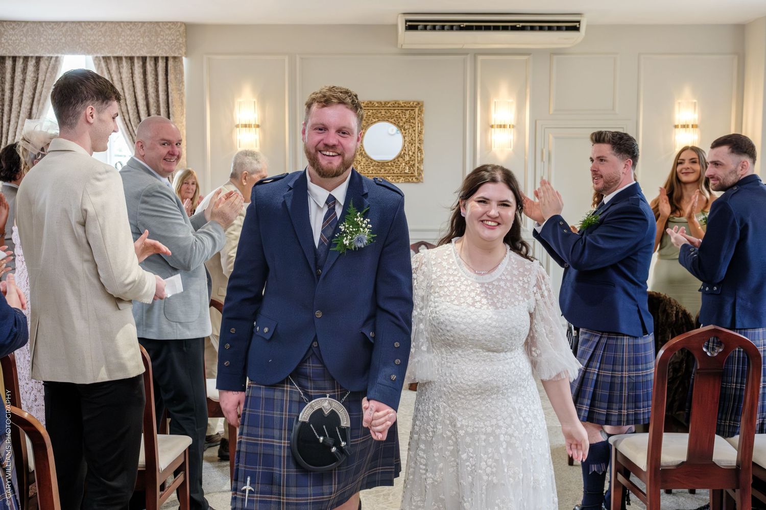 Happy newlyweds walk hand in hand after Scottish wedding ceremony, groom in tartan kilt, bride in white lace dress.