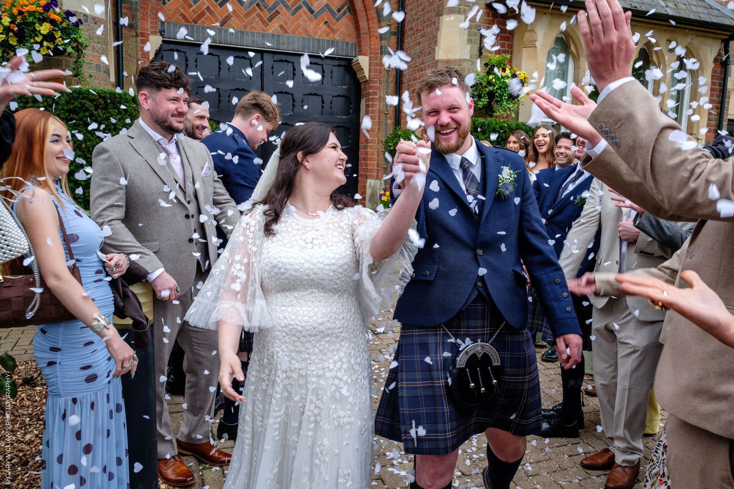 Bride and groom walk through confetti shower outside church, groom wearing blue kilt, surrounded by cheering wedding guests.