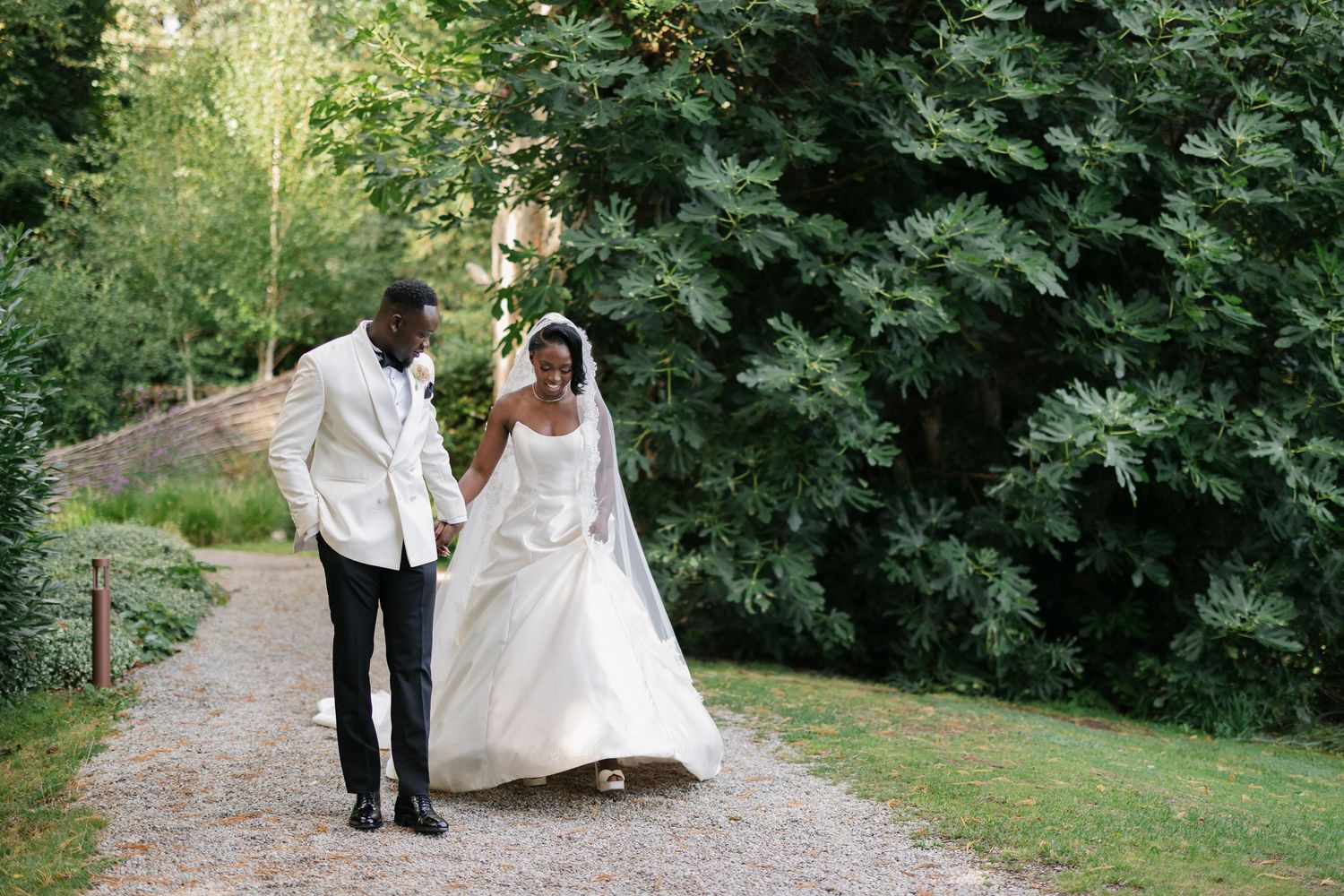 A bride and groom walk hand in hand along a garden path, she in a white gown, he in a white jacket.