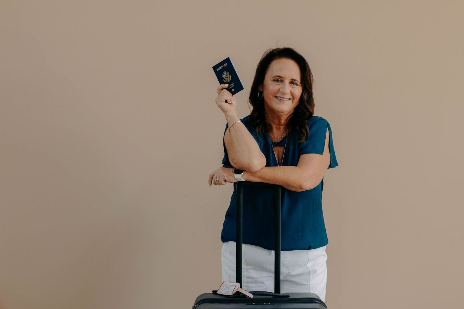 Woman in blue top leaning on luggage holding passport, ready for travel, isolated on beige background.