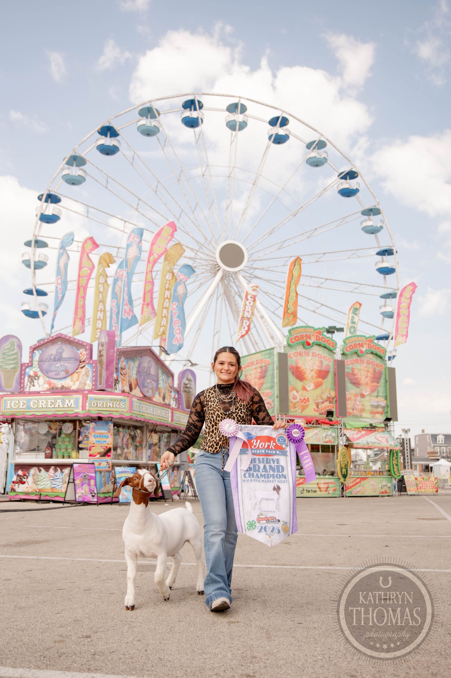Girl holding grand champion ribbon with goat at state fair, large Ferris wheel and colorful carnival rides in background.