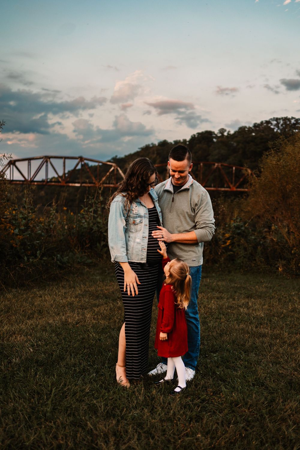 Pregnant woman with husband and young daughter in red dress pose outdoors near bridge at golden hour.