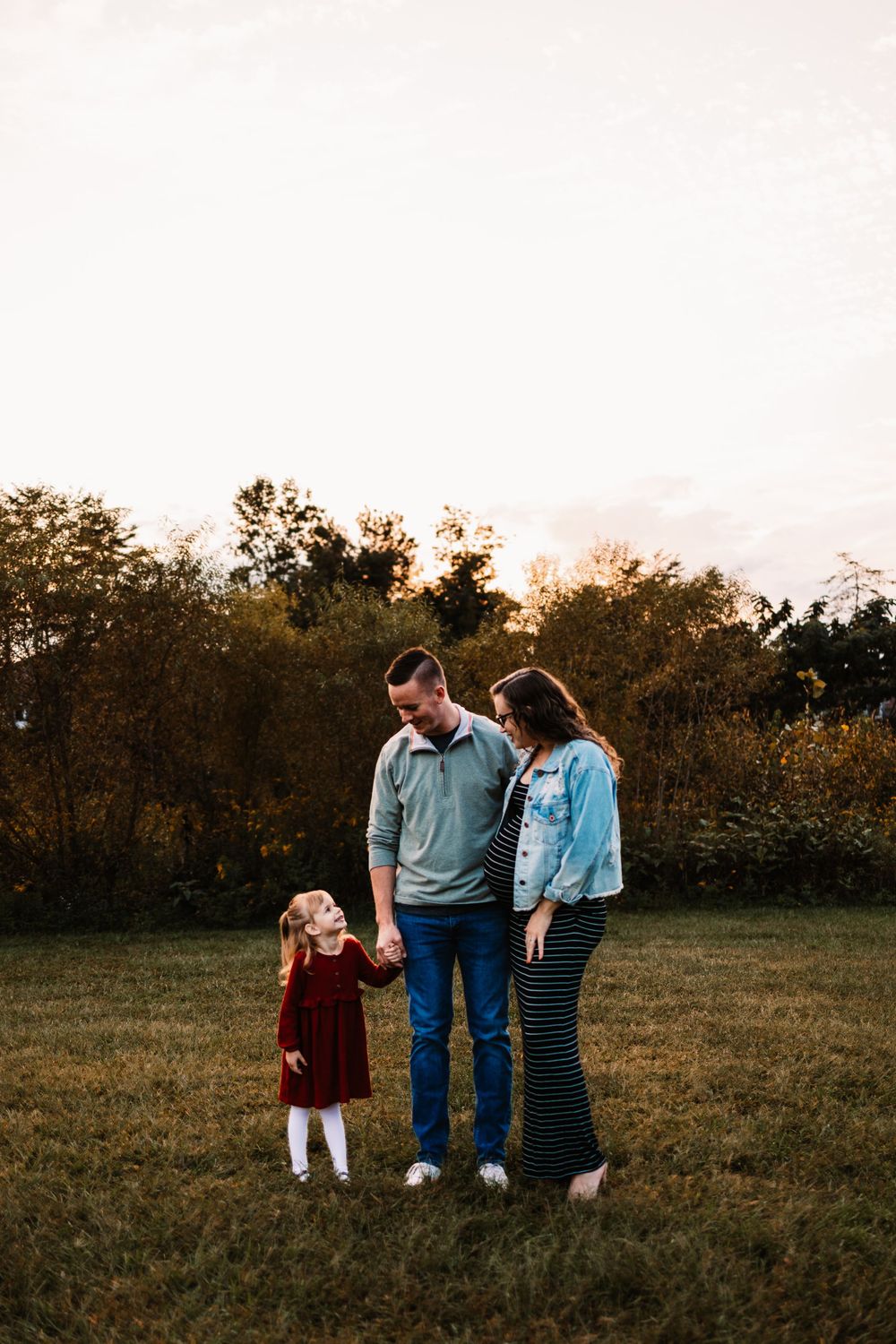A family of three stands in a golden field at sunset, parents holding hands with their young daughter in a red dress.
