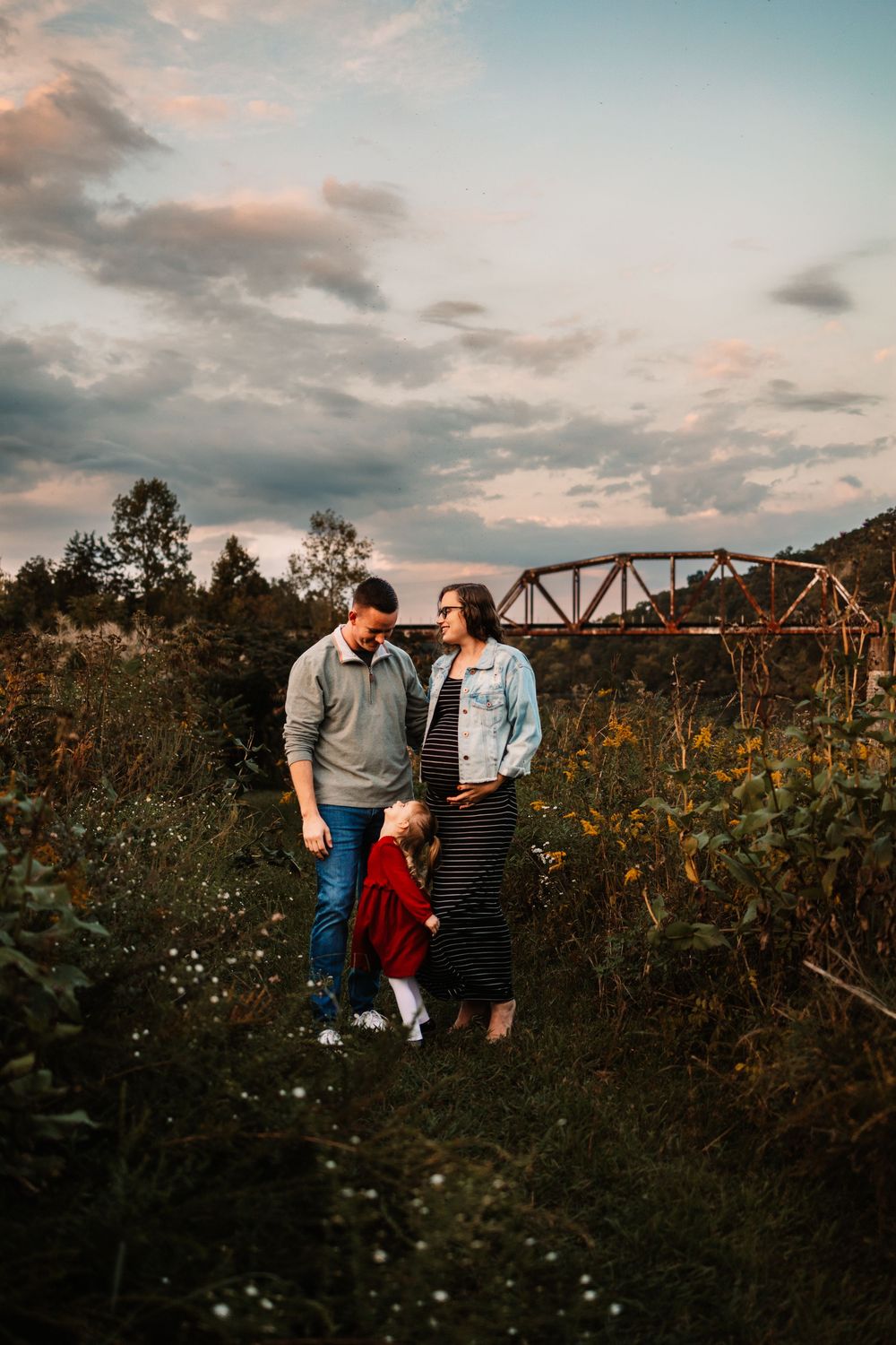 A family of three stands in a field at sunset, with a red bridge and lush greenery in the background.