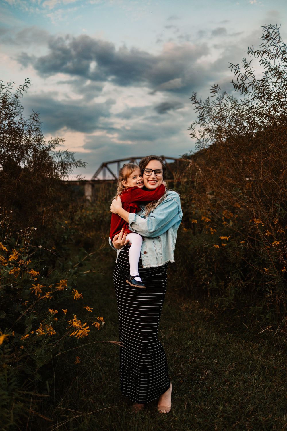 A smiling woman in glasses holds a toddler in a red sweater outdoors near yellow wildflowers and a bridge.