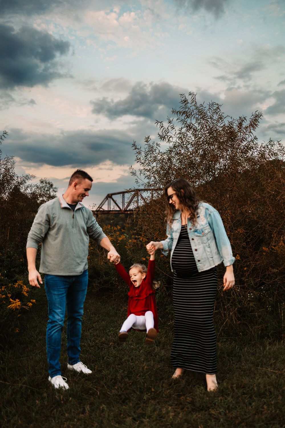 Pregnant woman and man swing toddler girl between them outdoors in autumn field at dusk.