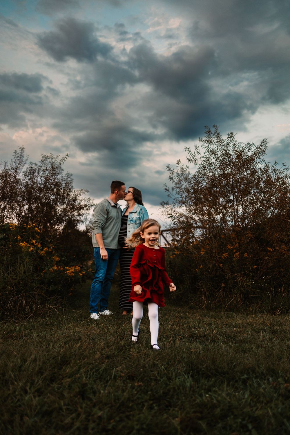 A young girl in a red dress runs outdoors while her parents share a kiss behind her under a dramatic cloudy sky.