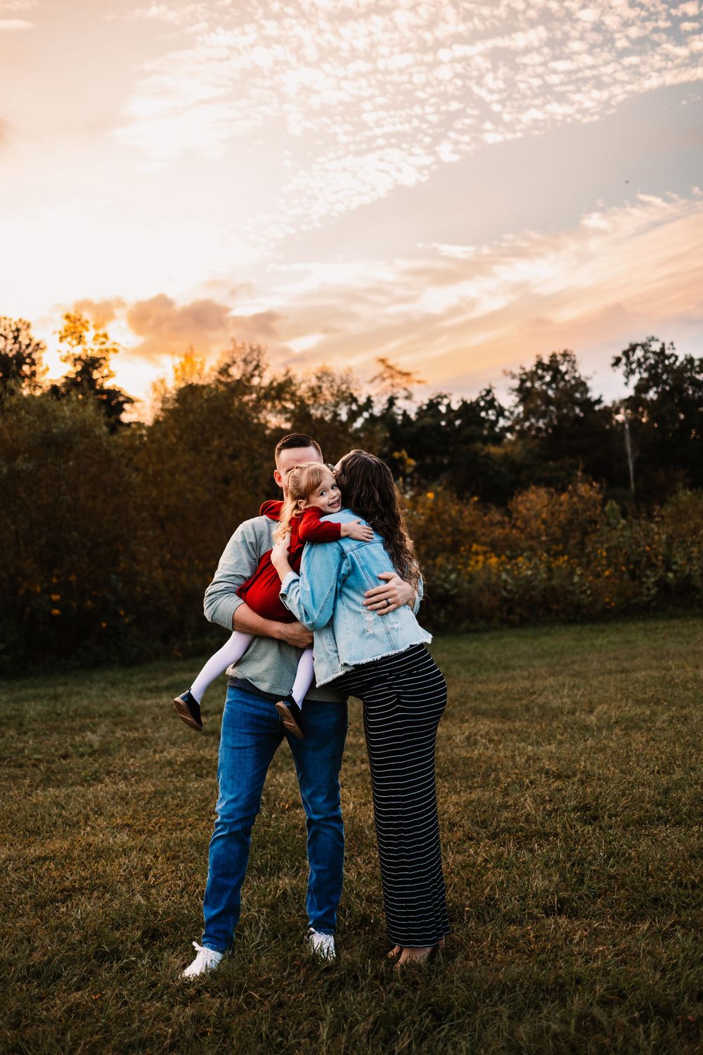 A couple kisses while holding their young child in a grassy field at sunset with colorful autumn trees in the background.