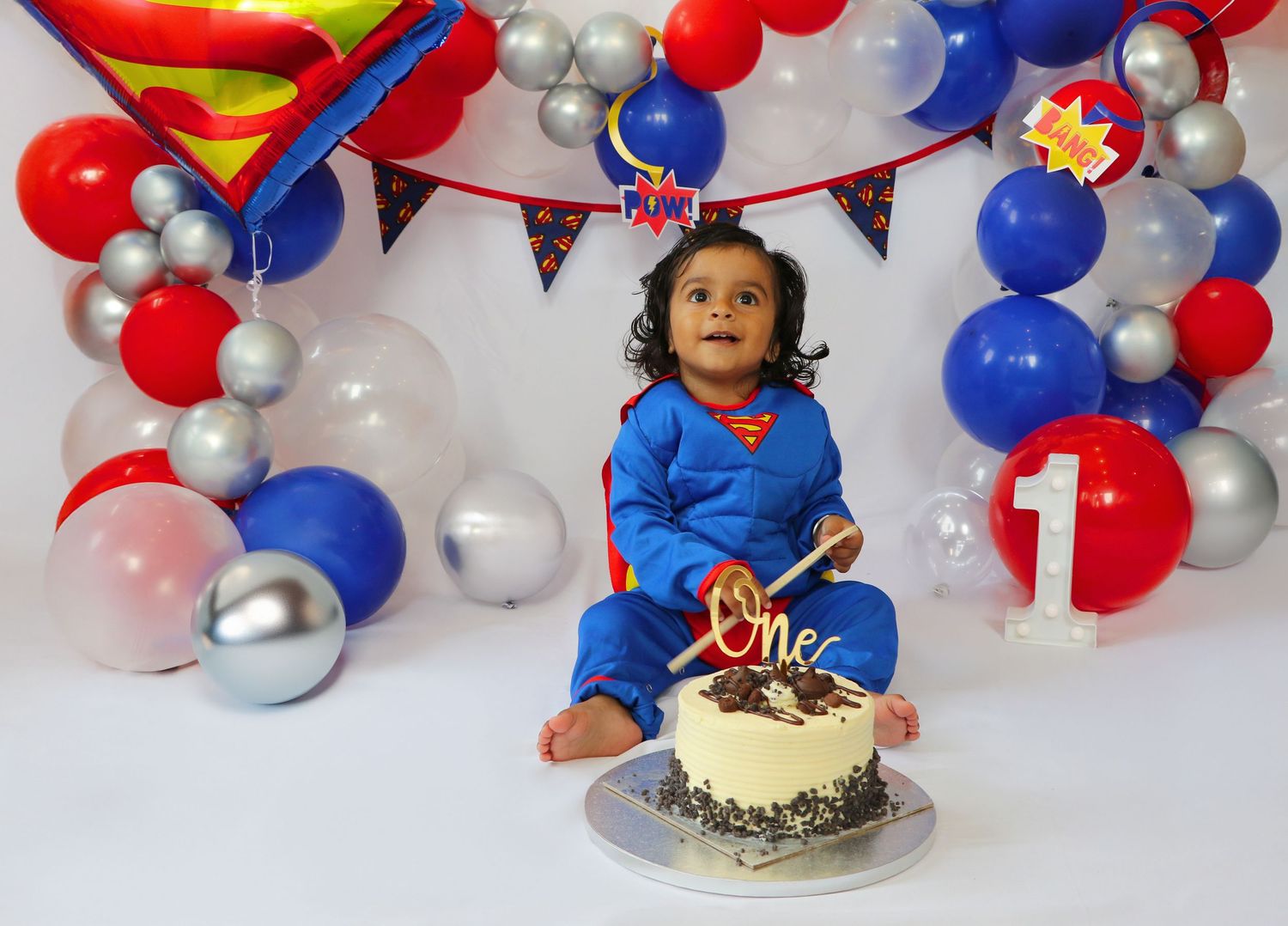 Baby in Superman outfit celebrates first birthday with cake, surrounded by red, blue, and silver balloons.