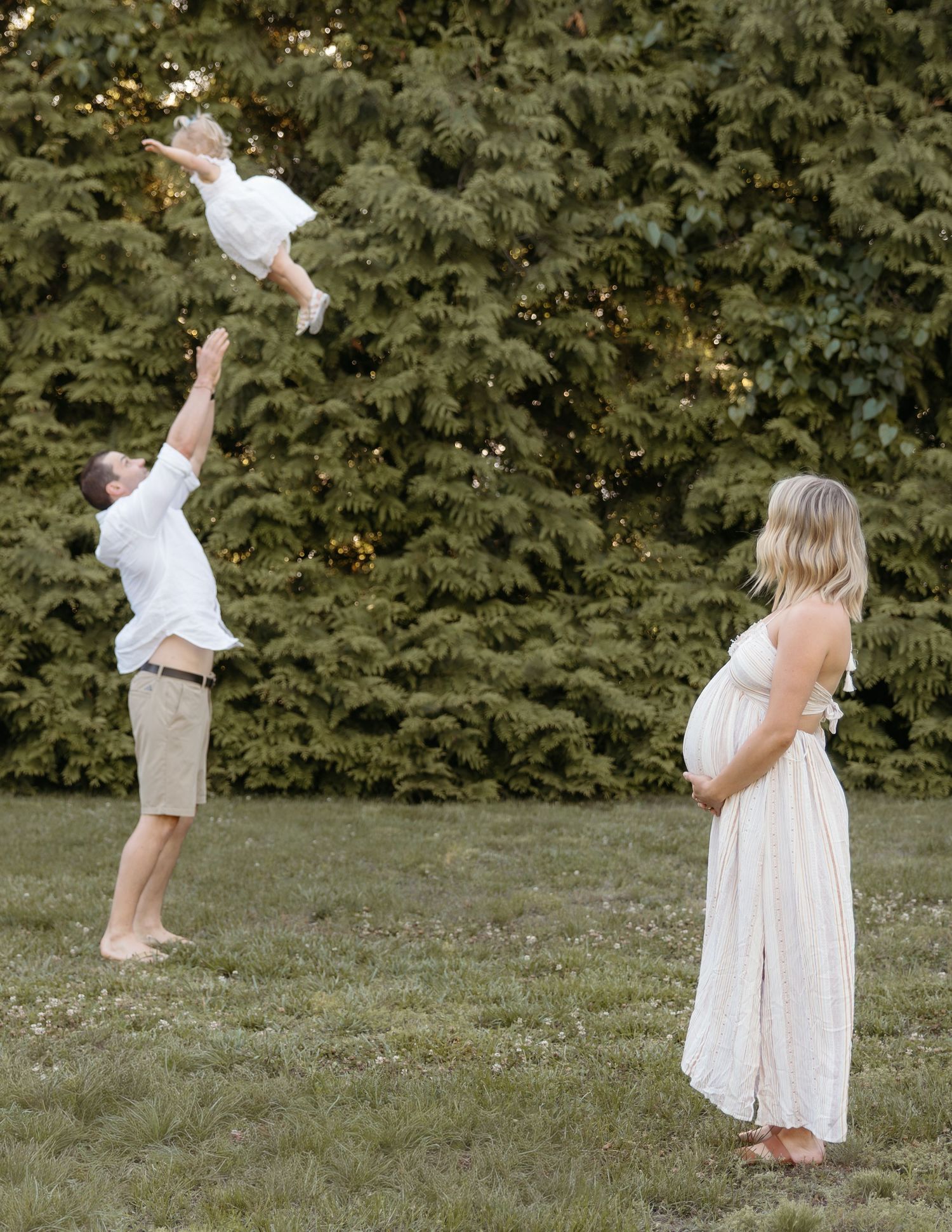 Pregnant woman in white dress watches man toss toddler in air during outdoor maternity photo session.