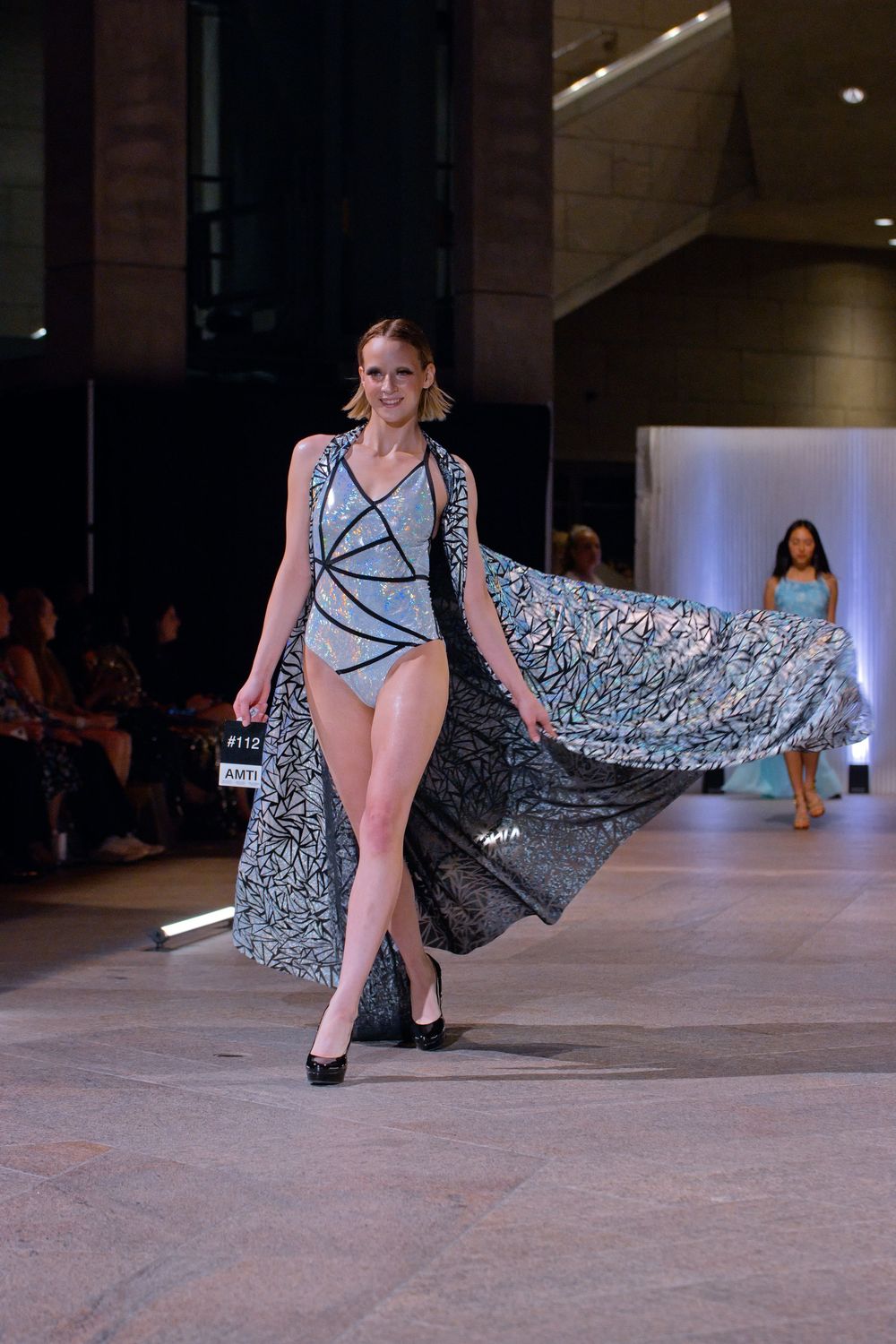 Model walks runway in blue patterned swimsuit with flowing sheer overlay cape at fashion show.