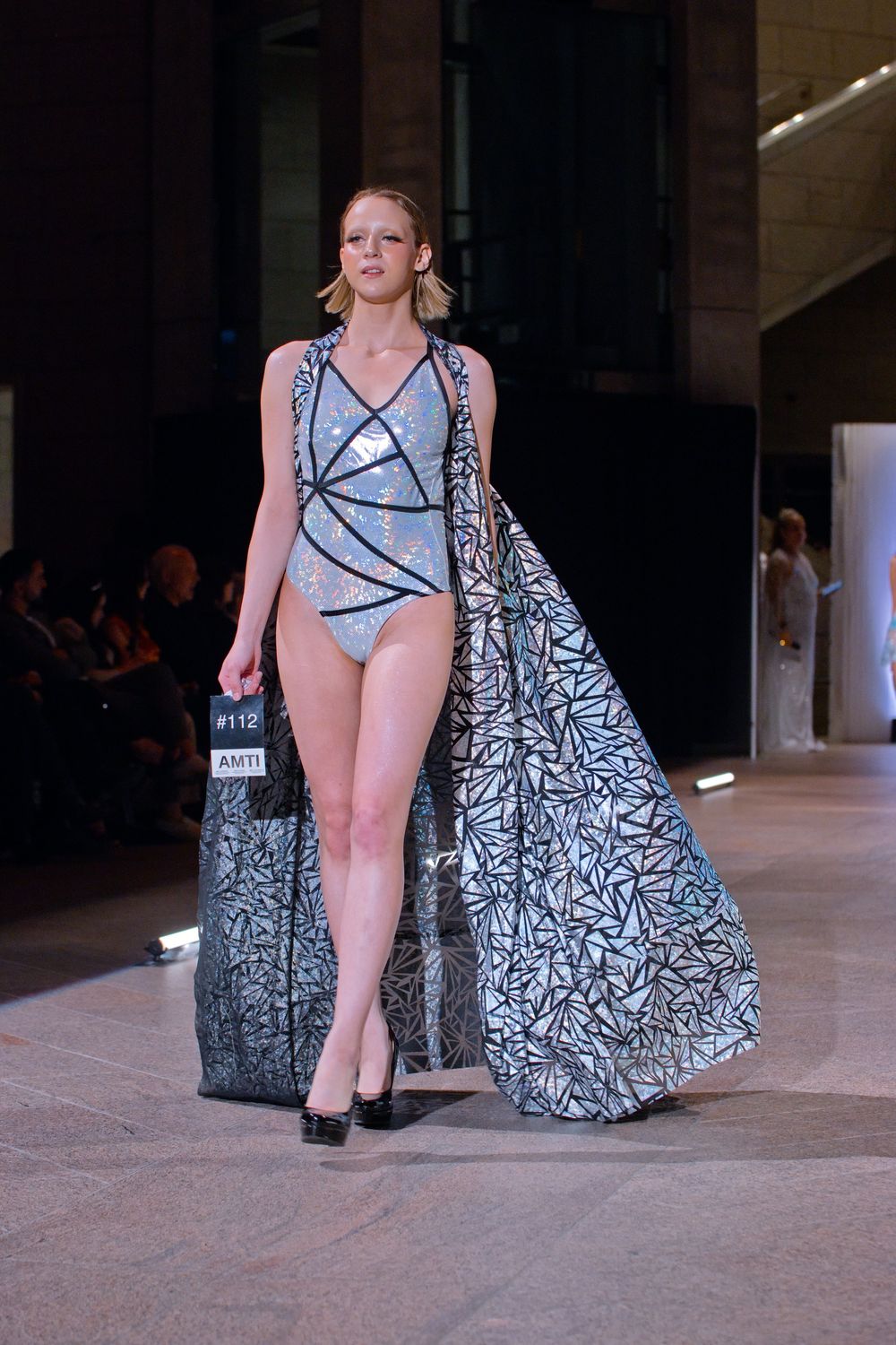 Model walks runway in silver sequined bodysuit with geometric patterned sheer duster coat at fashion show.