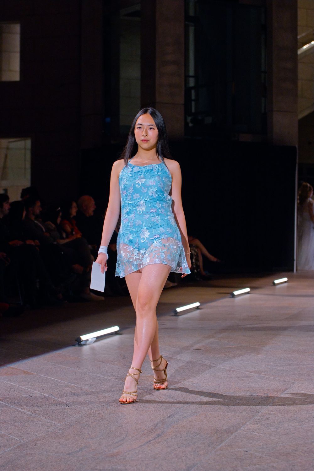 Asian female model walking runway in blue floral mini dress with strappy heels at fashion show.