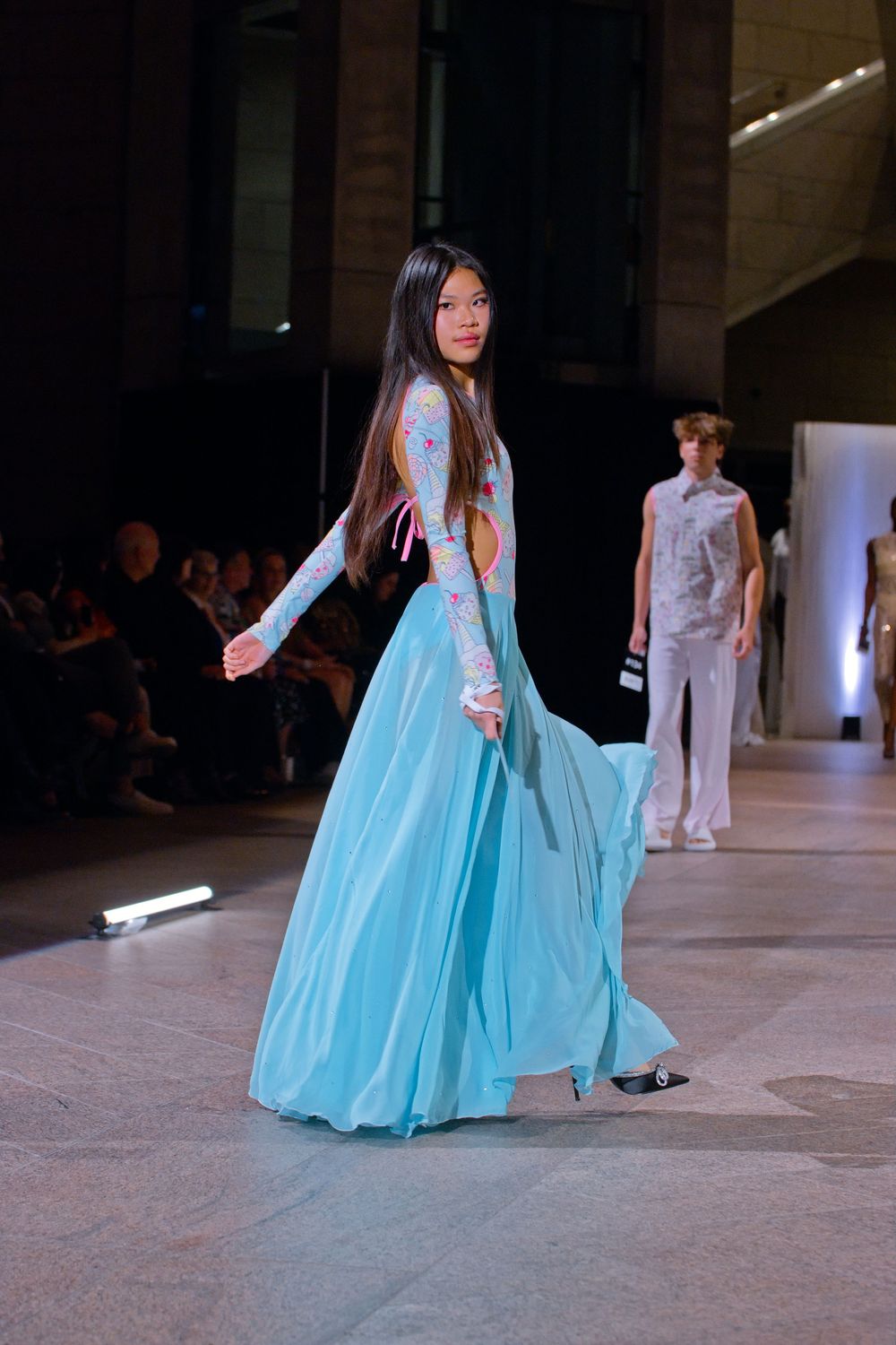 Model walks runway in a flowing turquoise gown with floral embroidered bodice at a fashion show.