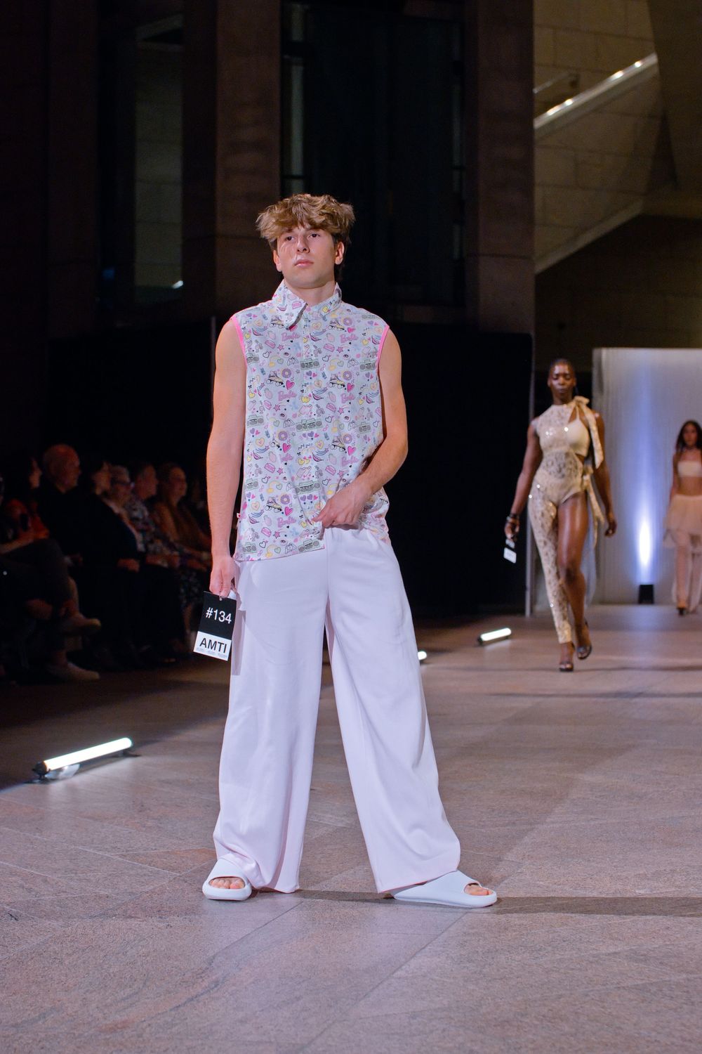 Male model walks runway in floral sleeveless shirt and wide-leg white pants with slide sandals at fashion show.