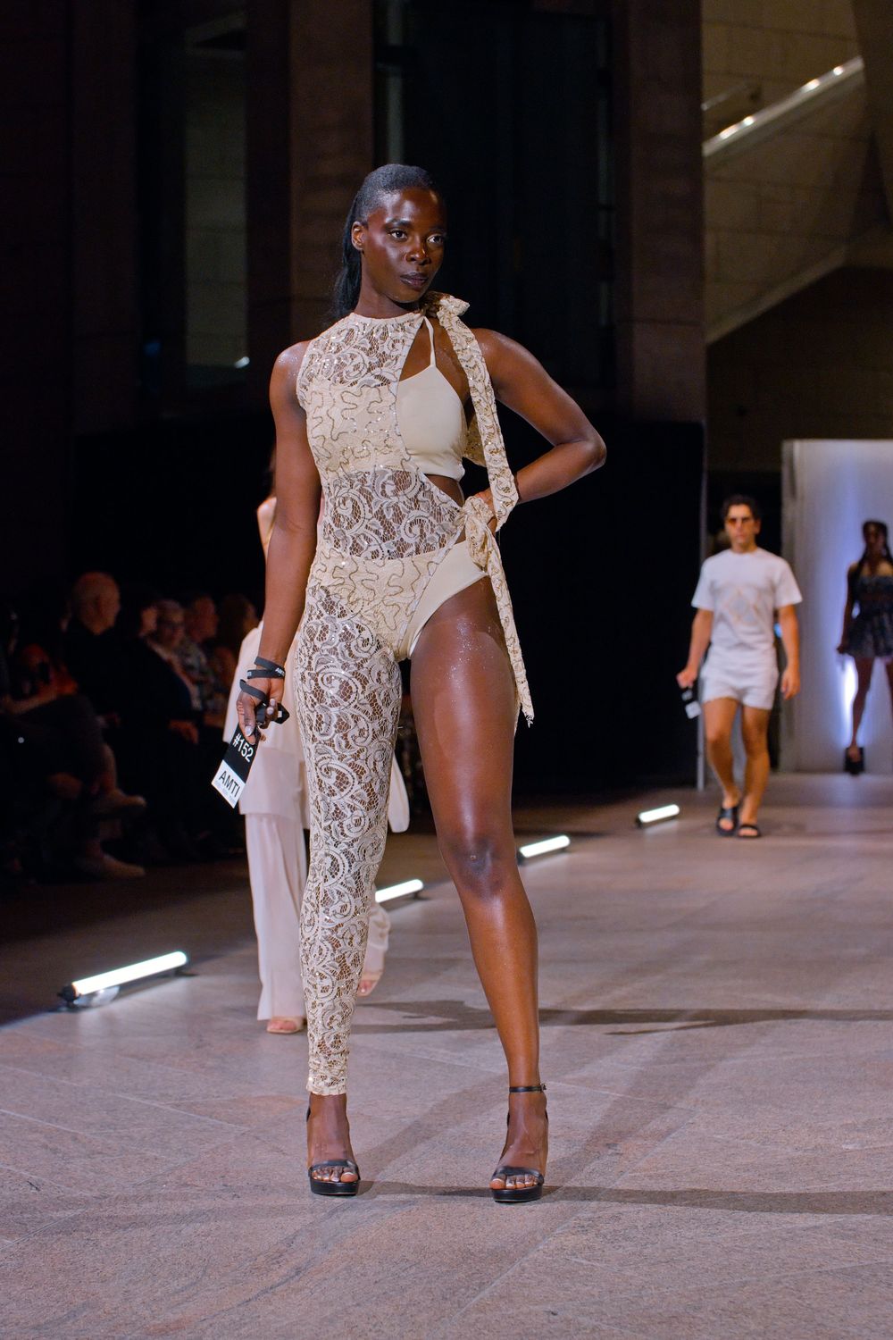 Model walks runway in cream lace cutout ensemble with sheer pants at fashion show.