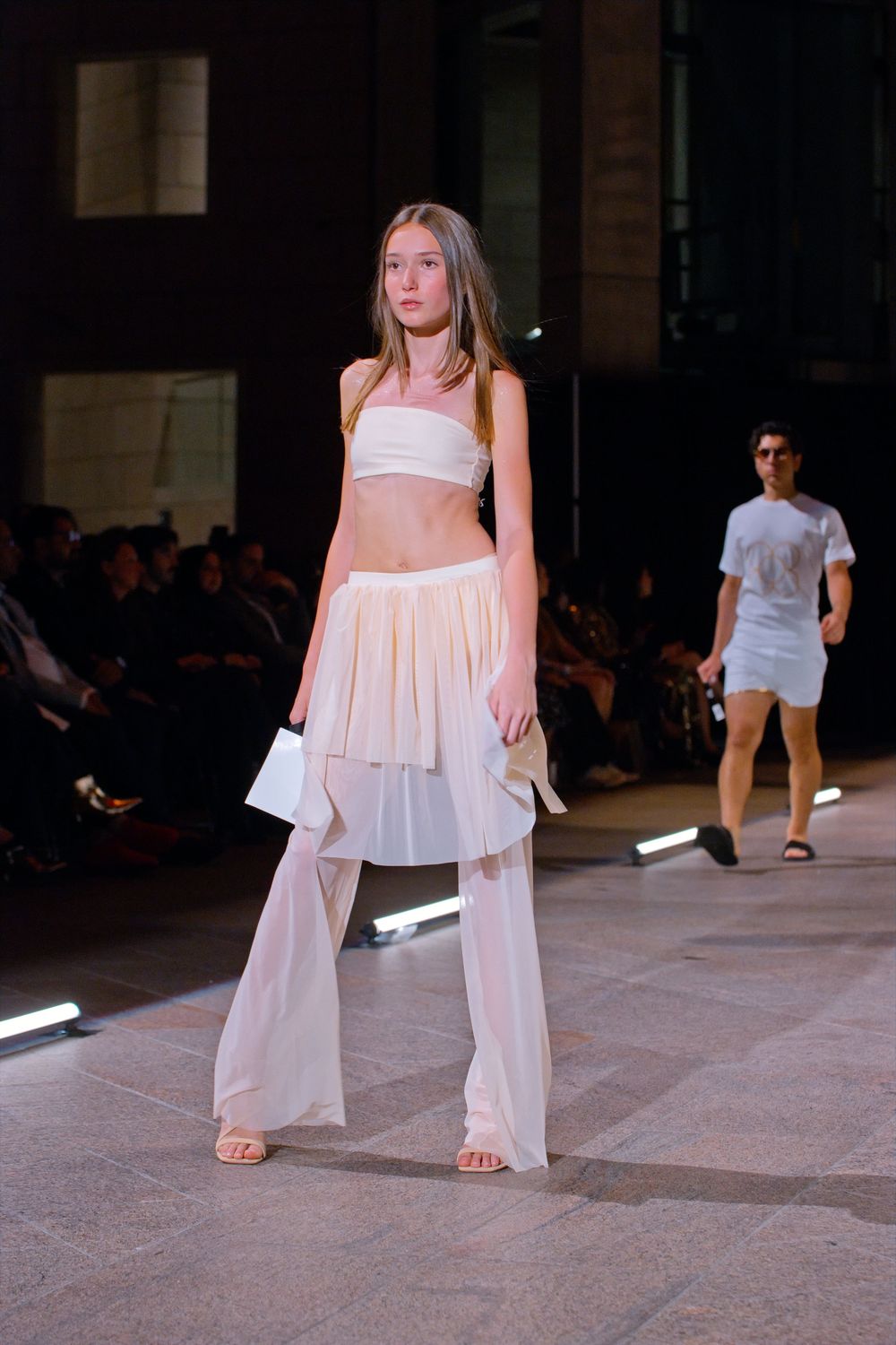 Model walks runway in white bandeau top, pleated mini skirt, and sheer wide-leg pants at fashion show.