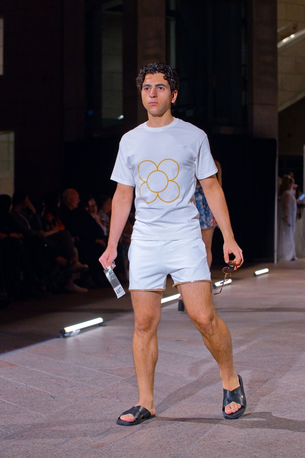 Male model walks runway in white graphic tee, shorts, and black slides at fashion show.