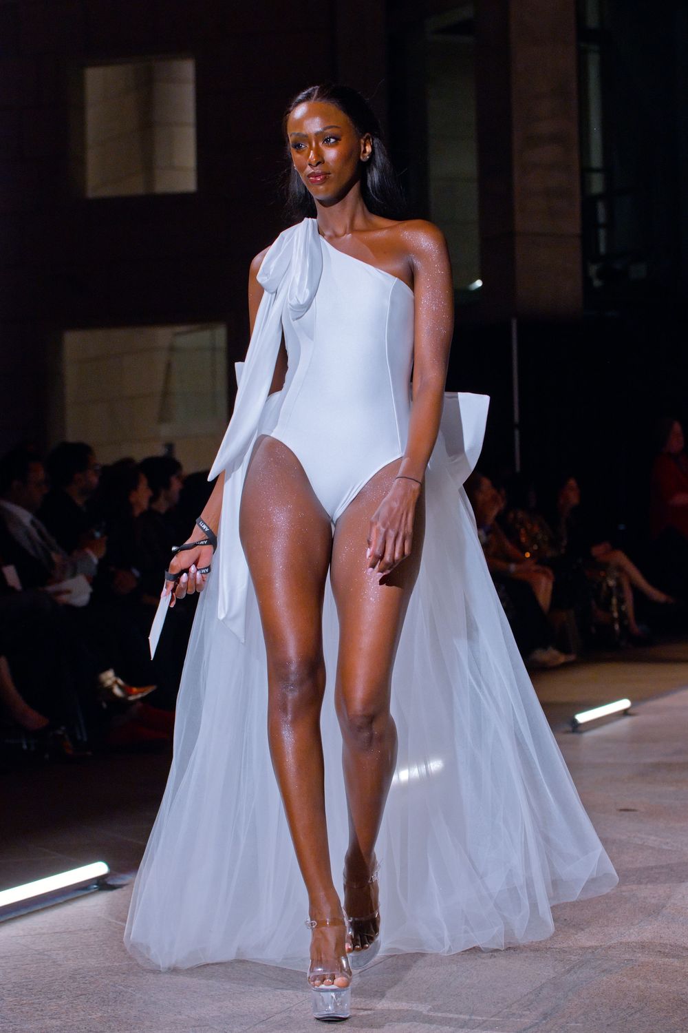 Female model walks runway in white one-shoulder bodysuit with flowing tulle skirt and bow detail at fashion show.