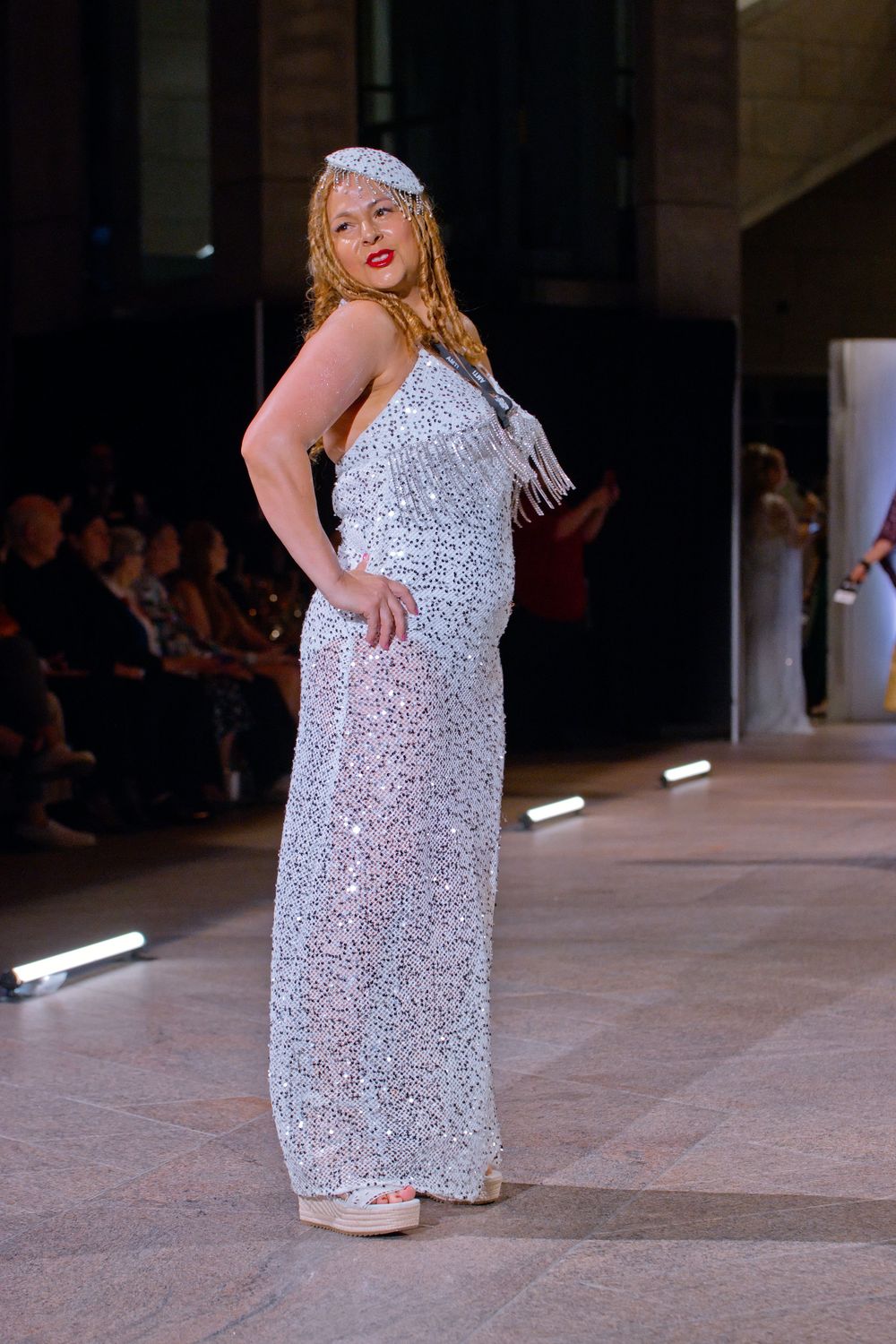 Model poses on runway wearing a full-length silver sequined gown with fringe headpiece at fashion show.