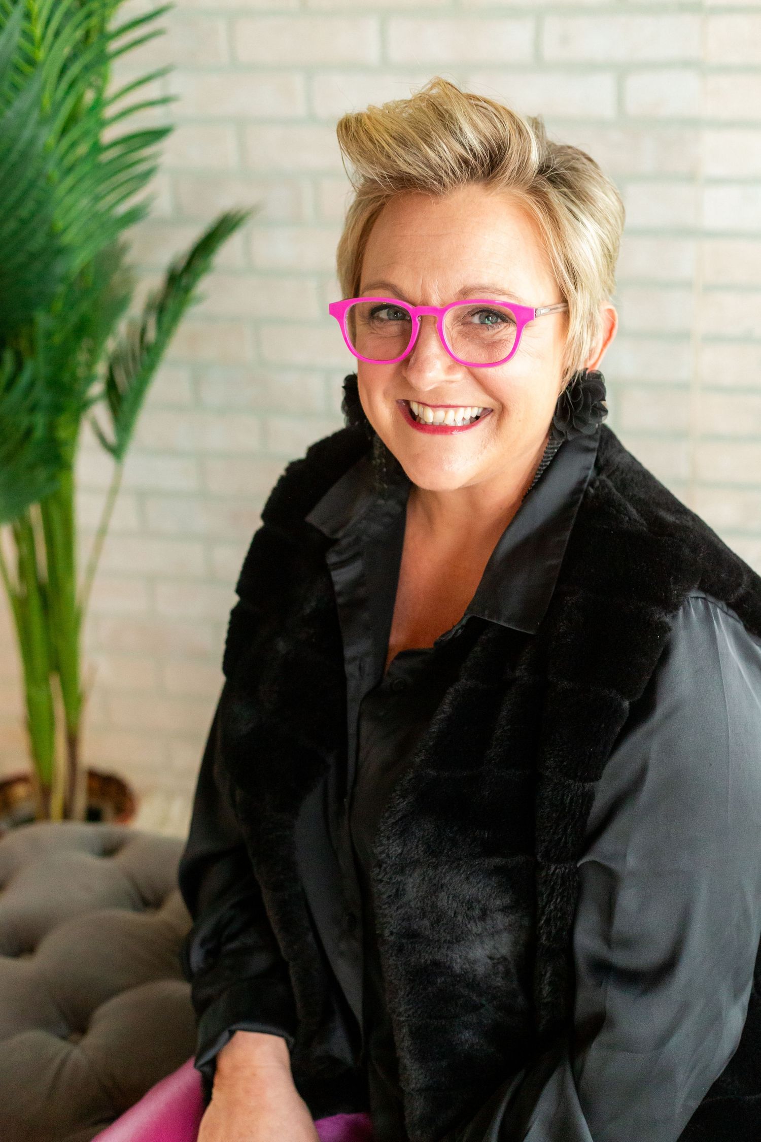 Smiling woman with short blonde hair, pink glasses, and black fur vest poses in a bright modern interior.