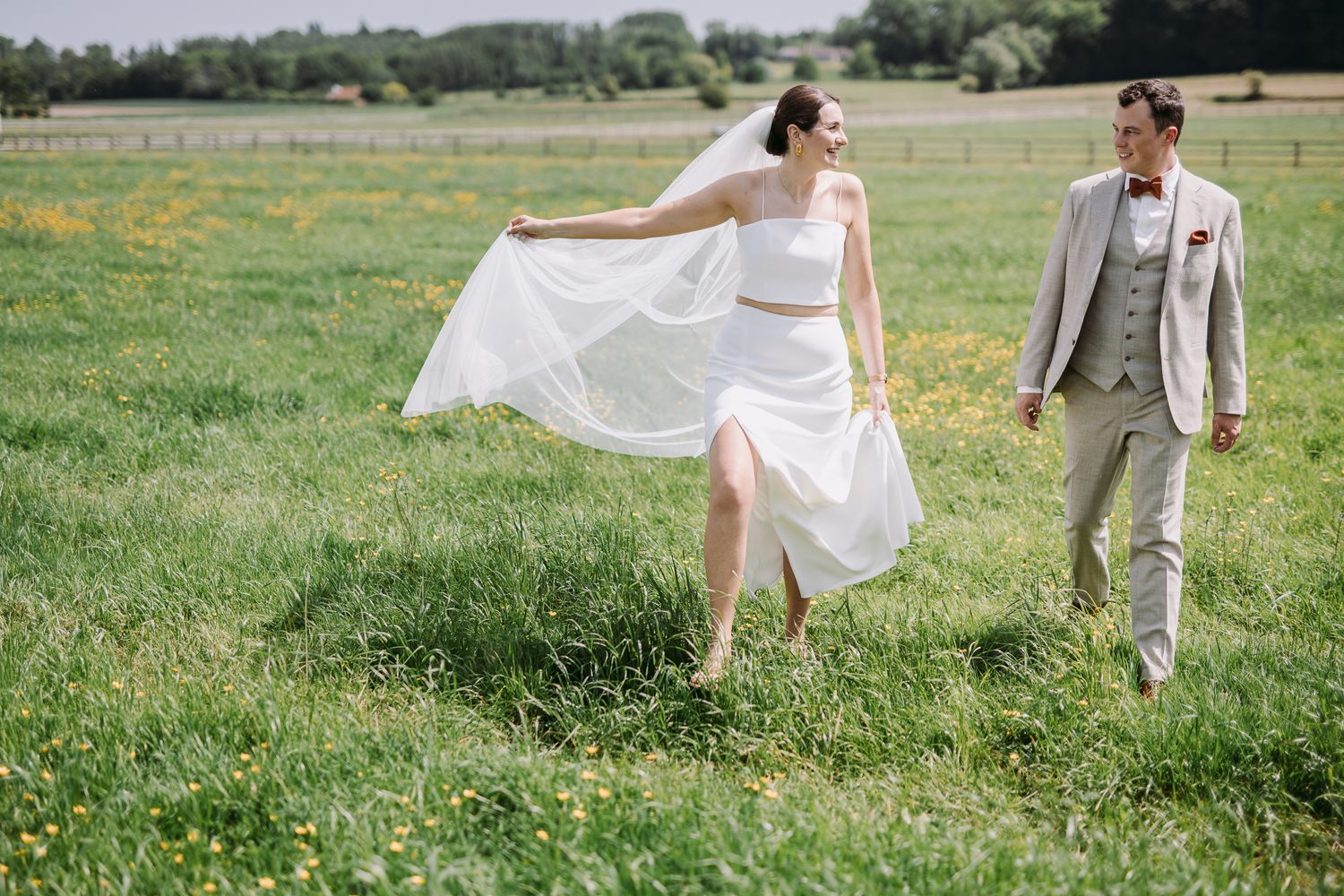 A bride and groom dance joyfully in a sunlit meadow, she in white crop top skirt, he in a light gray suit.