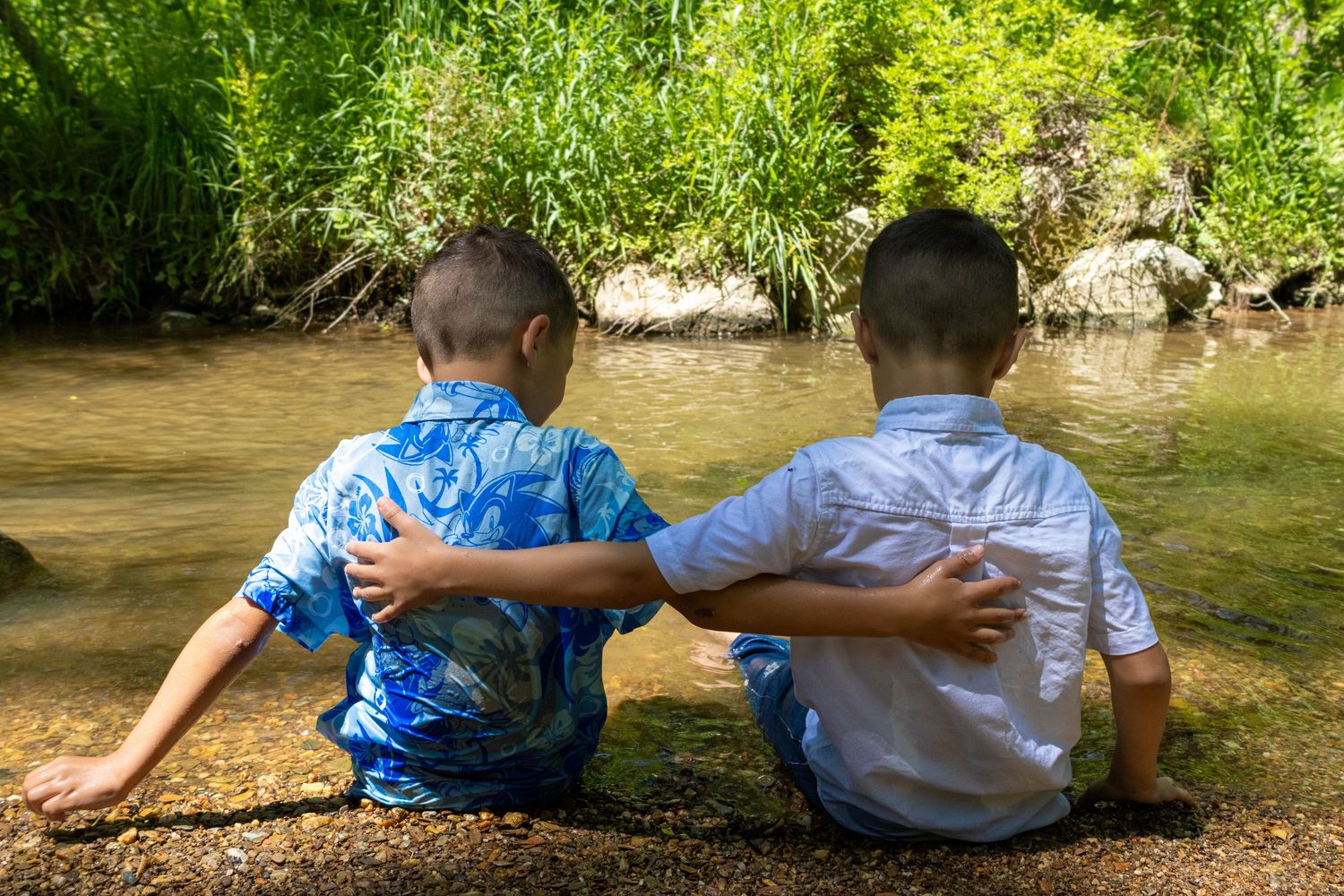 Two young boys sitting together by a peaceful stream, arms around each other, surrounded by lush green trees.