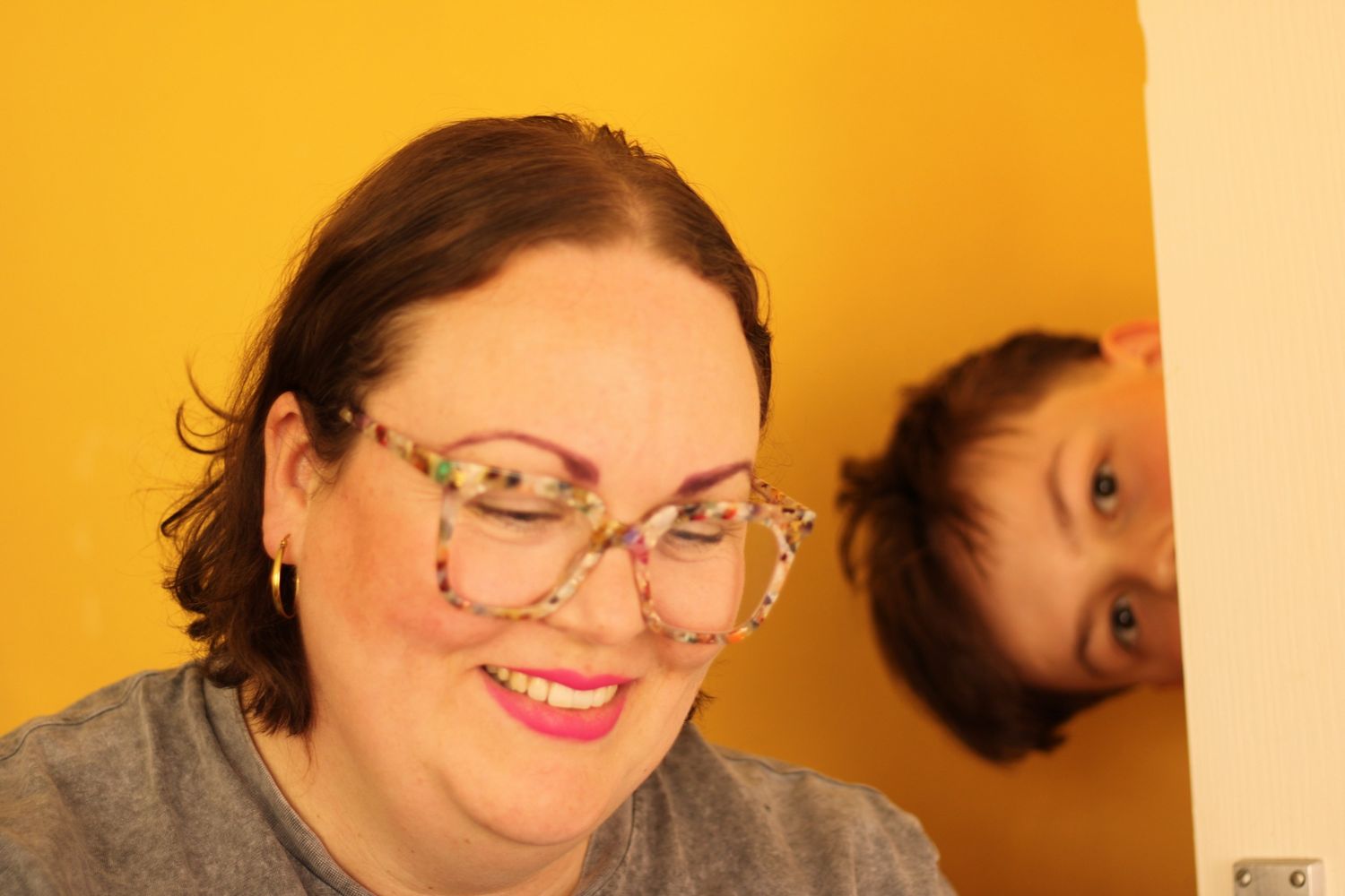 A smiling woman wearing decorative glasses with a child peeking from behind in front of a yellow wall.