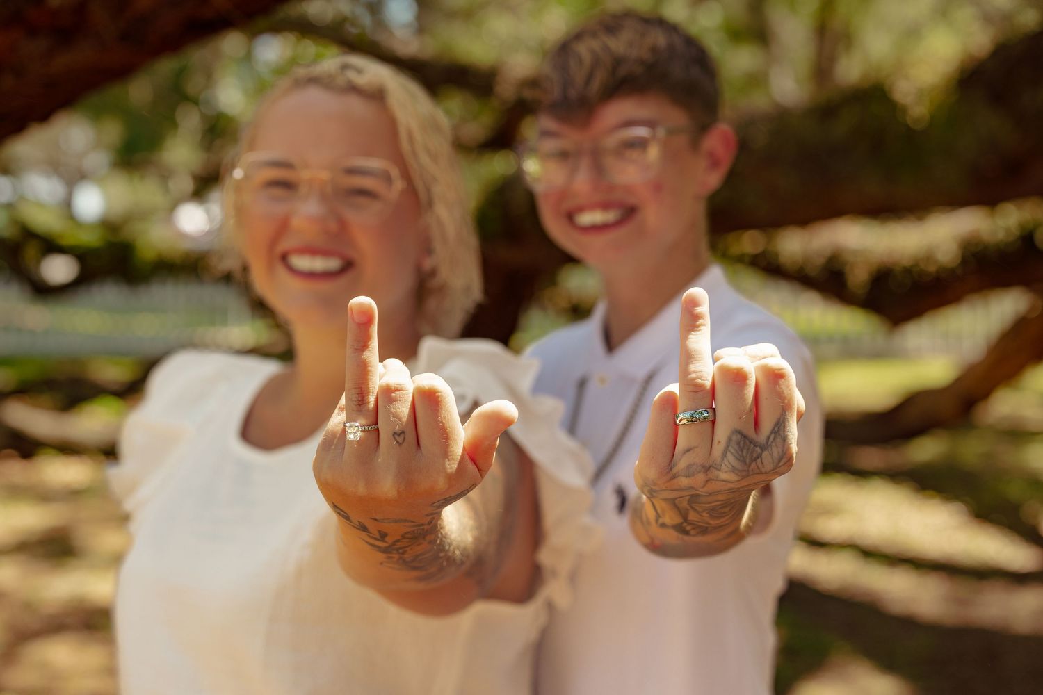 Two people showing off wedding rings with tattoos on their fingers, smiling outdoors under trees.