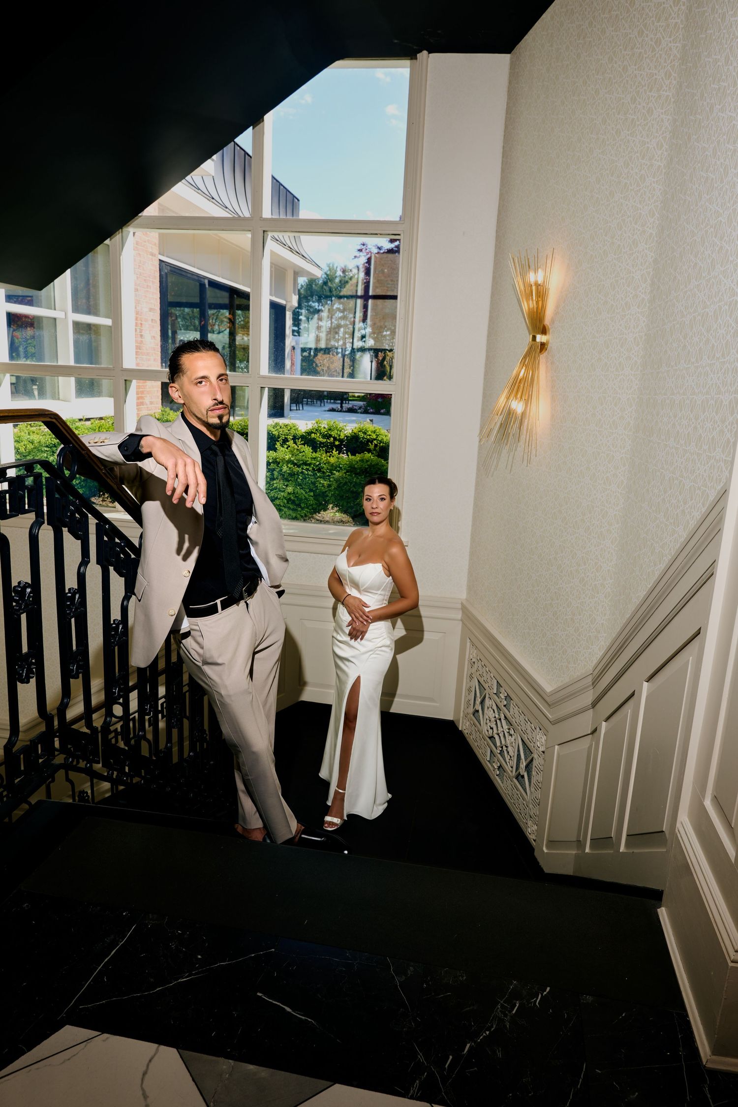 Elegant couple on grand staircase, groom in suit leads while bride in white satin gown poses in upscale hotel interior.
