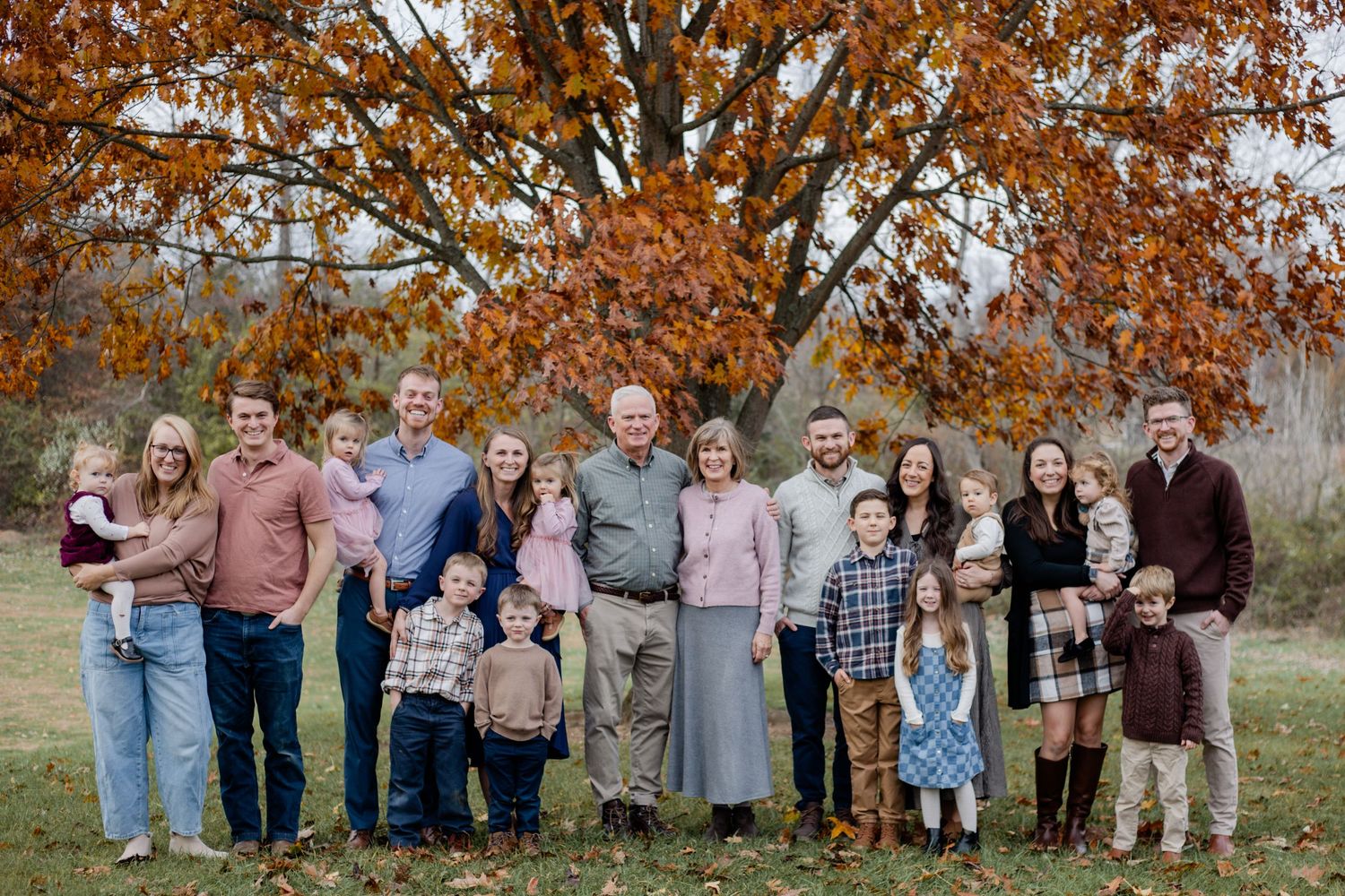 Large multigenerational family posing outdoors in autumn, with vibrant orange foliage and green lawn backdrop.