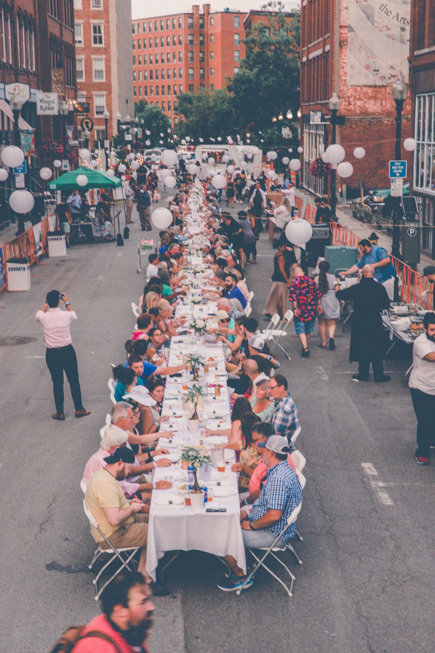 Aerial view of a long outdoor street dinner with crowds seated at white-clothed tables lined with decorations in an urban setting.