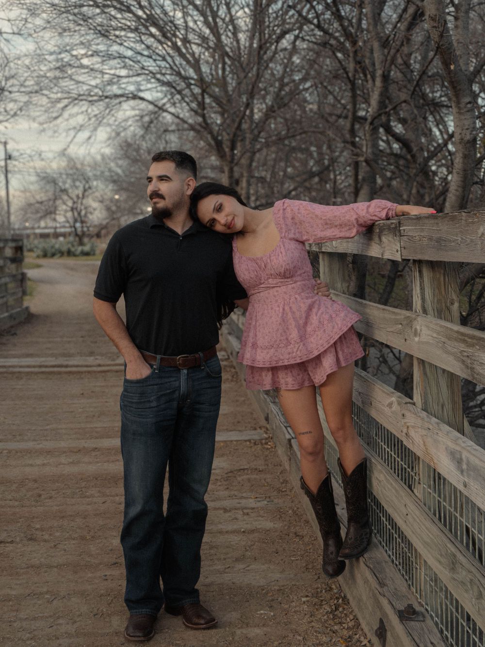 Couple posing by a rustic wooden fence, woman in pink dress and cowboy boots leaning on mans shoulder.