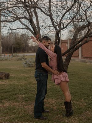 Couple leaning in for a kiss while standing beneath a bare tree on a grassy lawn at dusk.