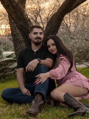 Engaged couple sitting together under a tree at sunset, woman displaying diamond engagement ring.