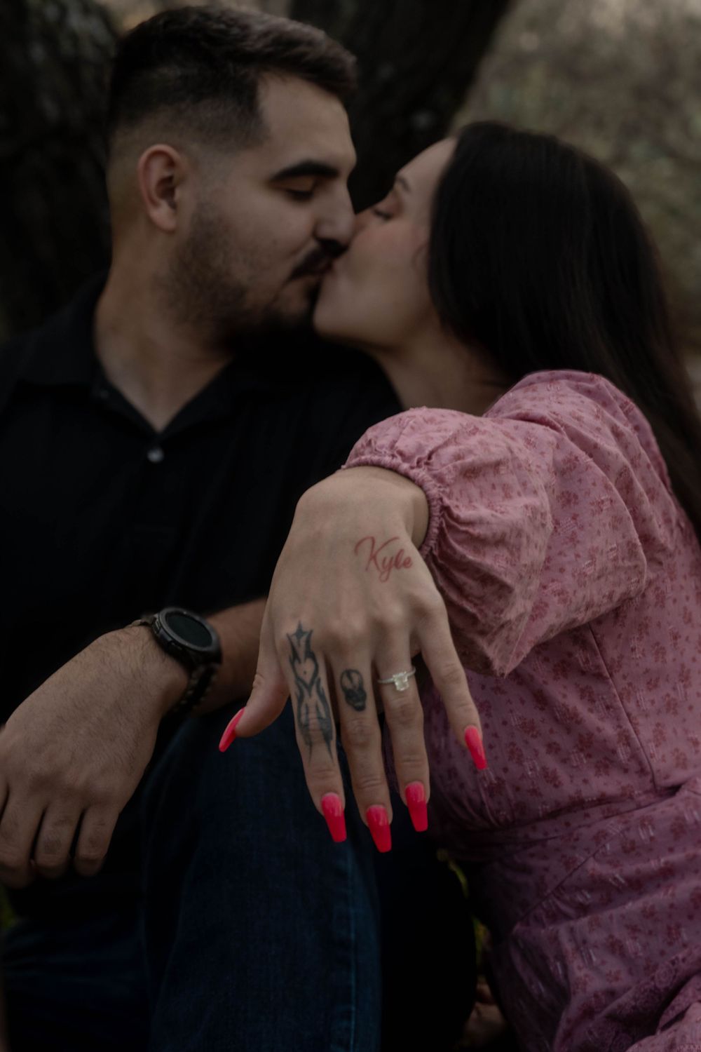 Engaged couple kissing while woman displays diamond engagement ring and hand tattoos toward camera.