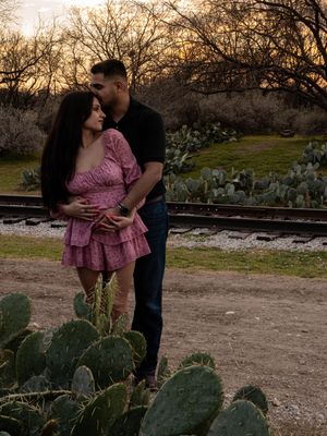 Romantic couple embracing near railroad tracks surrounded by prickly pear cactus at golden hour.