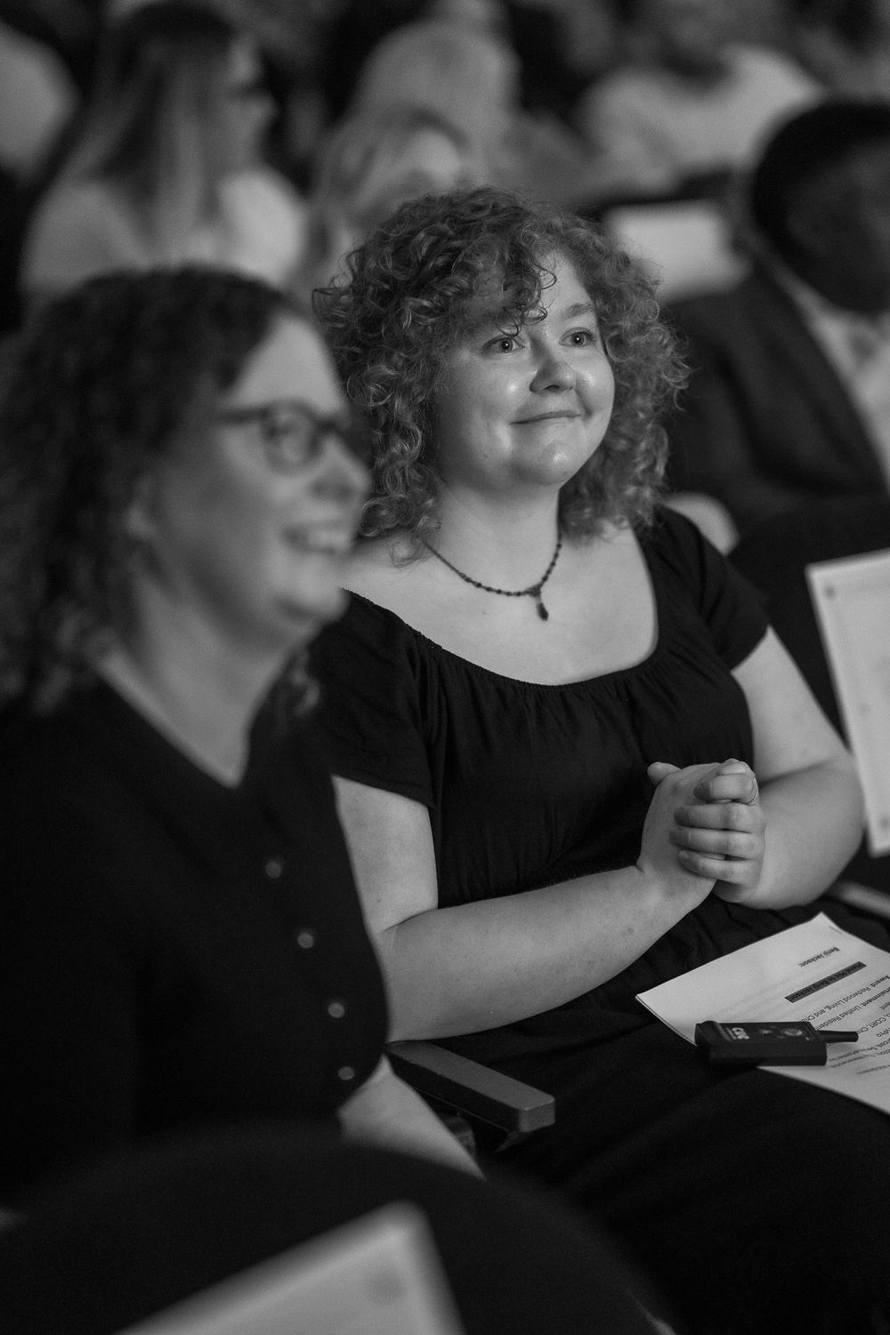 Two women smiling in an audience at an event, black and white photo, curly-haired woman in black dress with necklace.