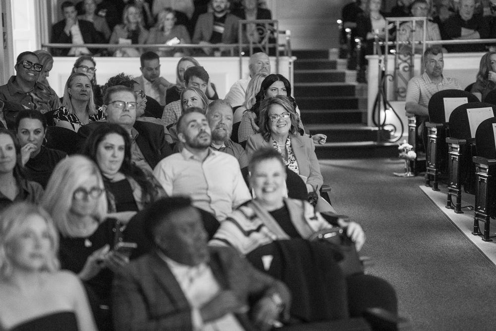 Black and white photo of a diverse audience seated in rows, smiling and engaged at an indoor event or show.