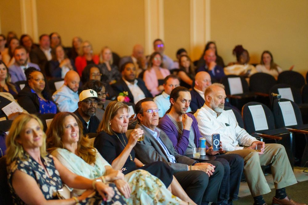A diverse audience attends an indoor event, seated in rows of chairs in an elegant venue.