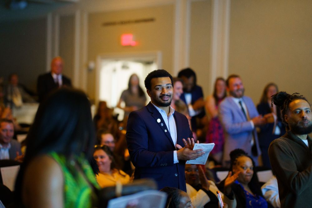 Man in navy blazer speaks at a formal indoor event with a diverse seated audience in an elegant venue.