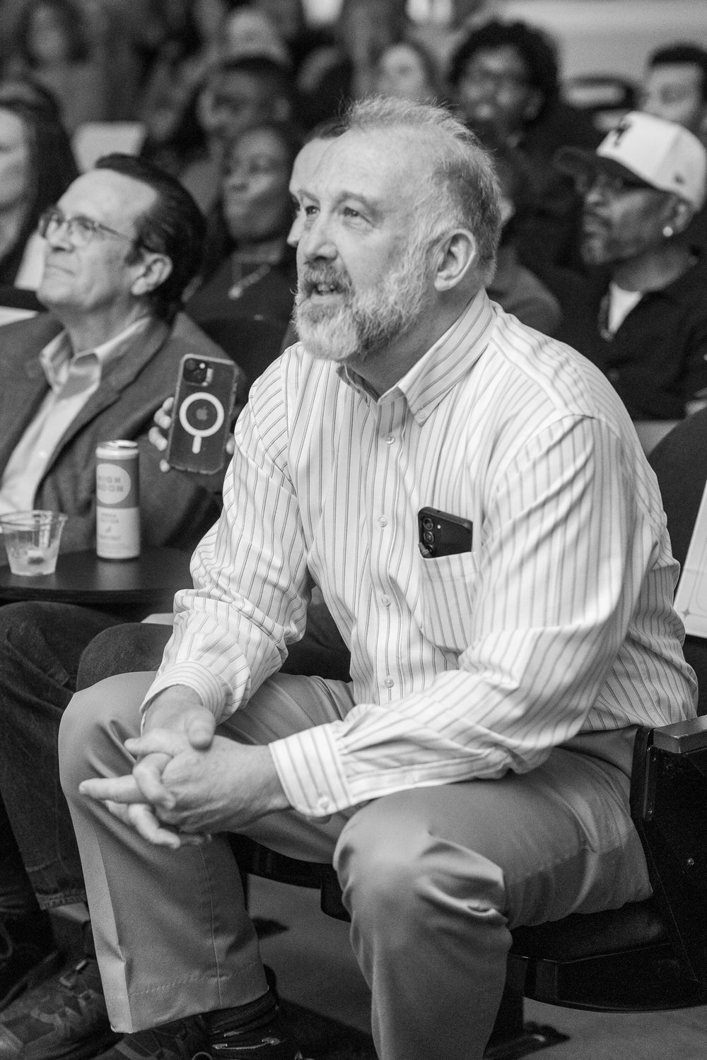 Black and white photo of a bearded man in a striped shirt sitting attentively in a crowded audience.