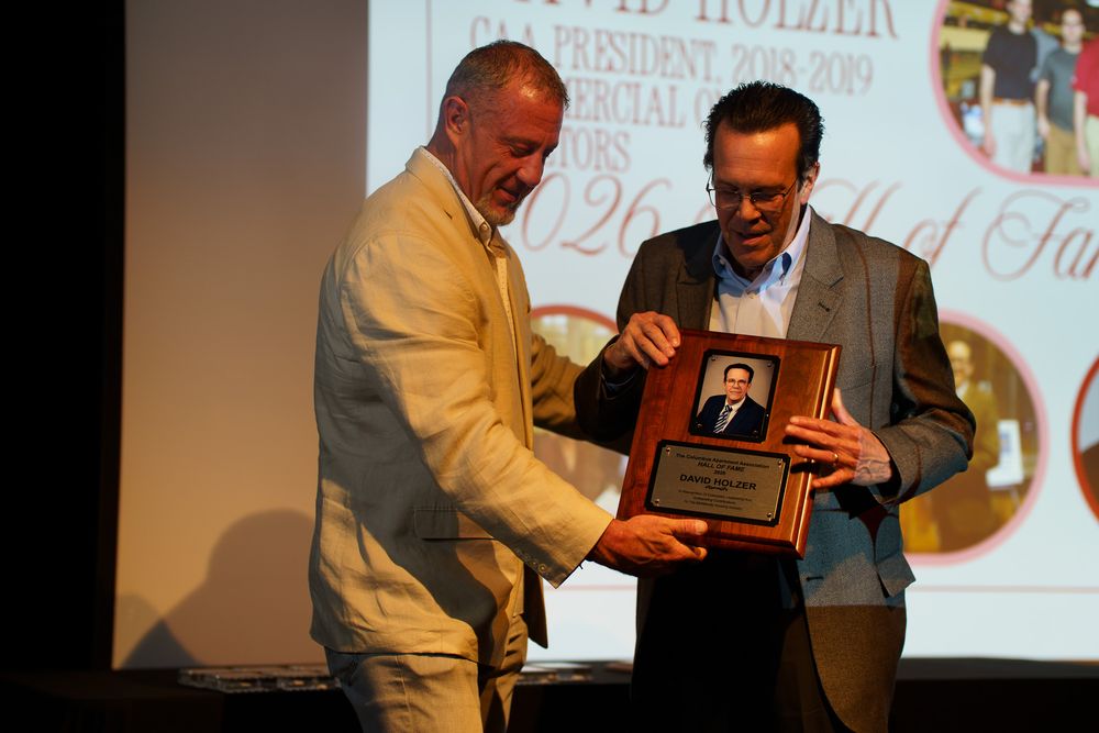 Two men on stage during an award ceremony, one presenting a plaque to the other at a formal recognition event.