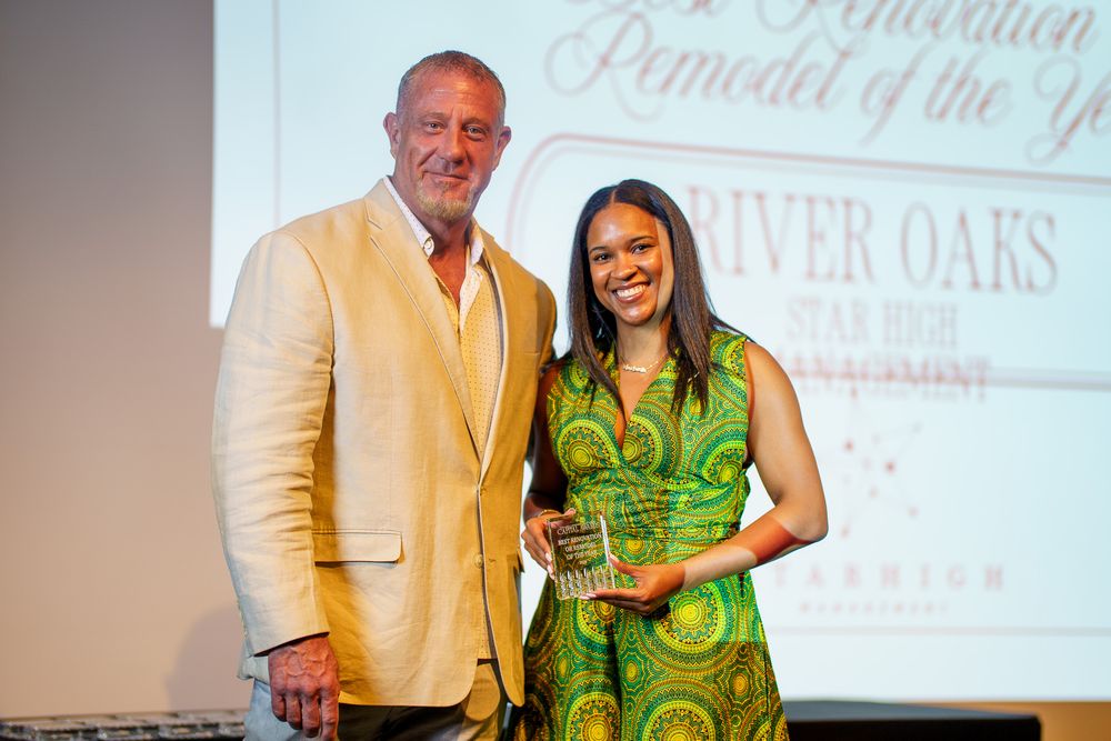 Two people pose together on stage at an awards ceremony, holding a glass trophy at River Oaks.