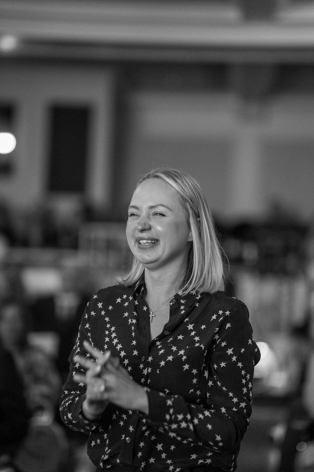 Black and white photo of a smiling blonde woman in a star-patterned blouse clapping at an indoor event.