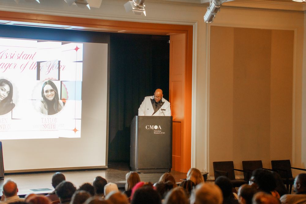 A speaker presents at a podium during a conference with a projected presentation screen in a modern auditorium.
